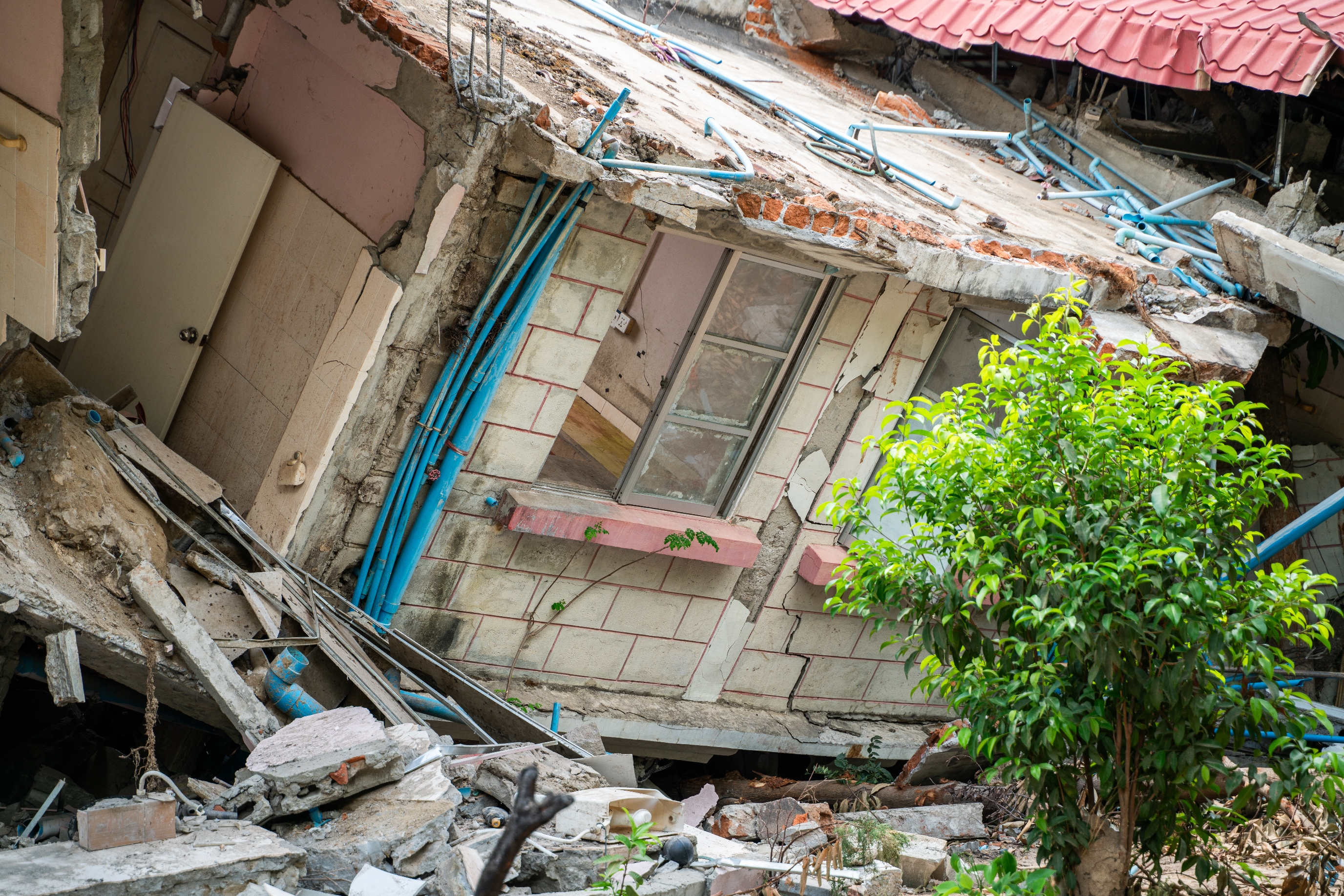 Collapsed building with visible plumbing and debris, tilted on its side. A small tree stands in front, seemingly undisturbed by the damage