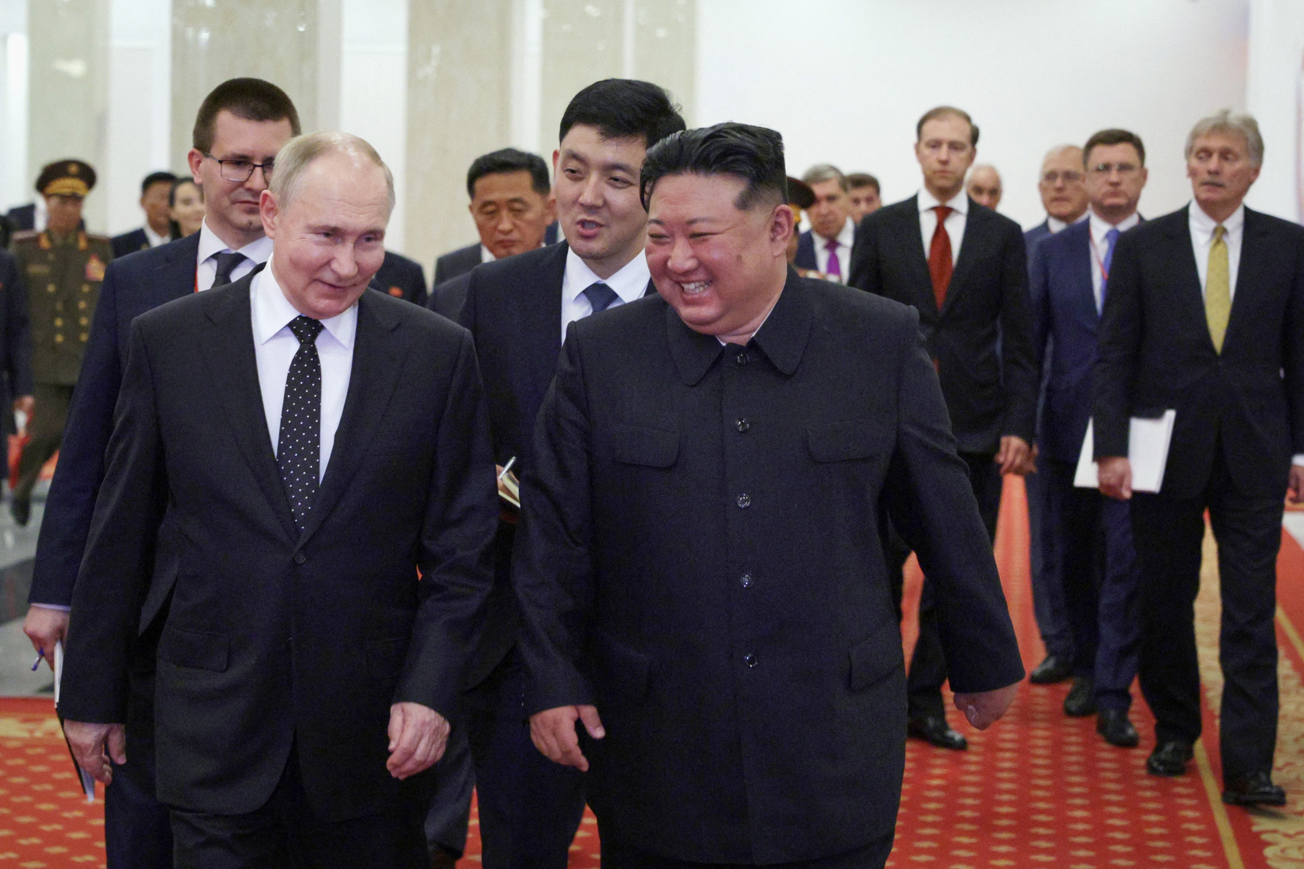 A group of officials, including two in suits, walk together on a red carpet, engaged in conversation amidst a formal setting