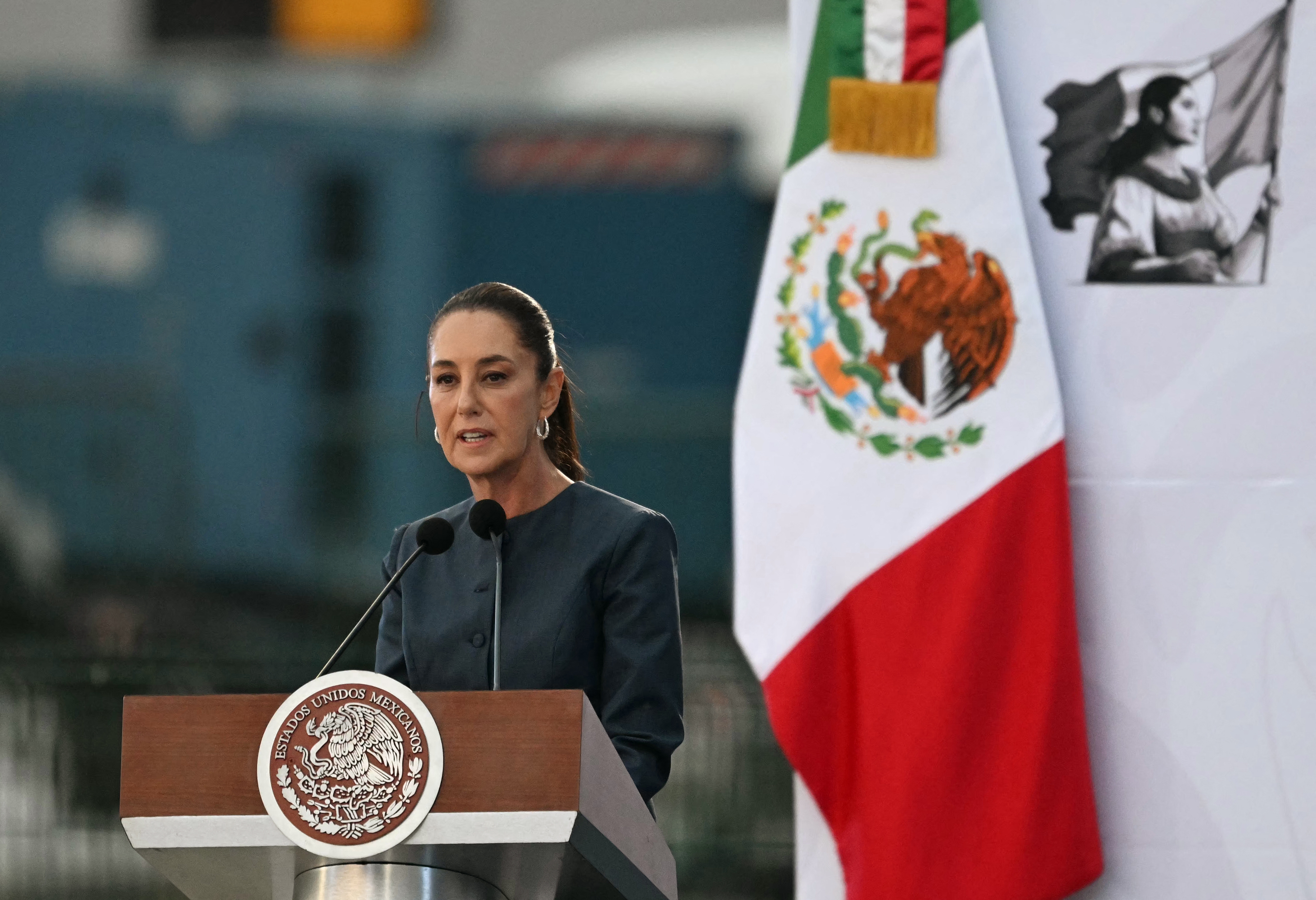 A woman speaks at a podium with the Mexican flag emblem. The Mexican flag and a historical image are seen in the background