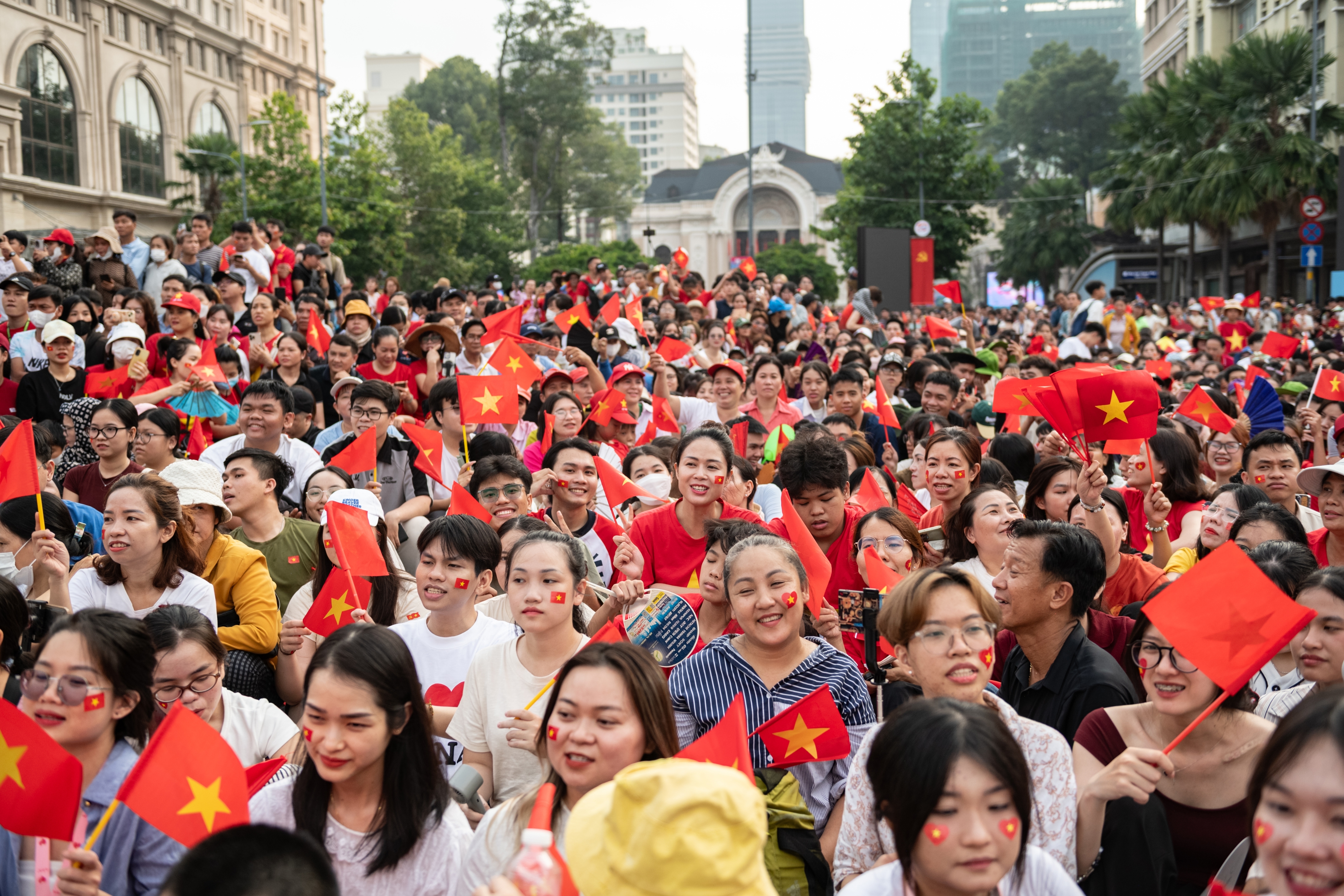 A large crowd celebrates in a public square, holding national flags and wearing festive attire, with visible excitement and joy on their faces