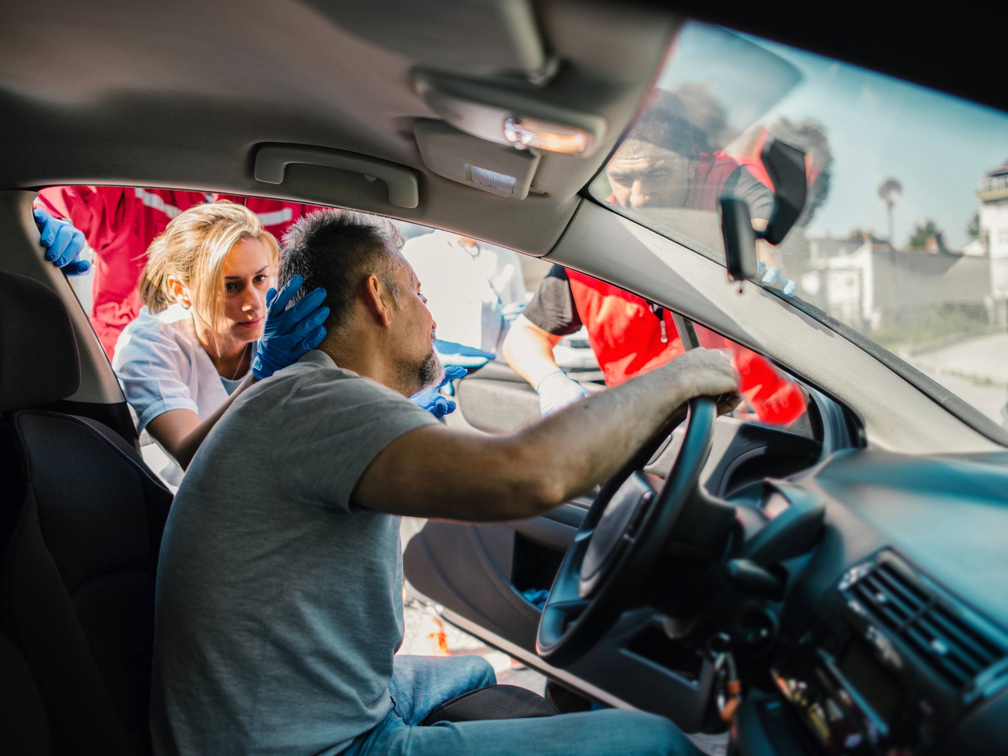Emergency responders assist an injured man sitting in a car after an accident, checking his head and neck for injuries