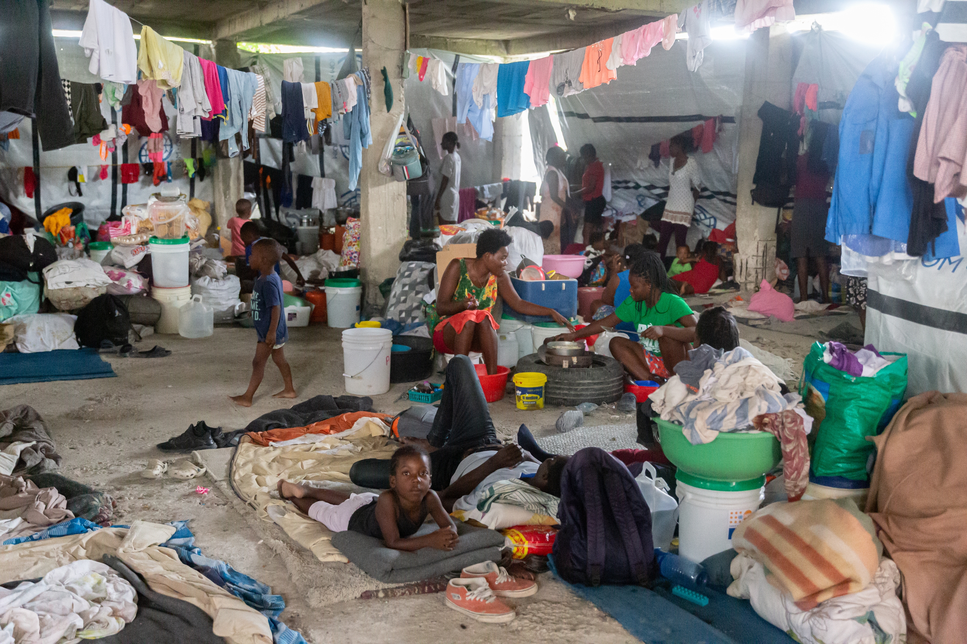 People in a makeshift shelter with clothing hanging, people sitting, lying on mats, surrounded by supplies, creating a sense of community and resilience