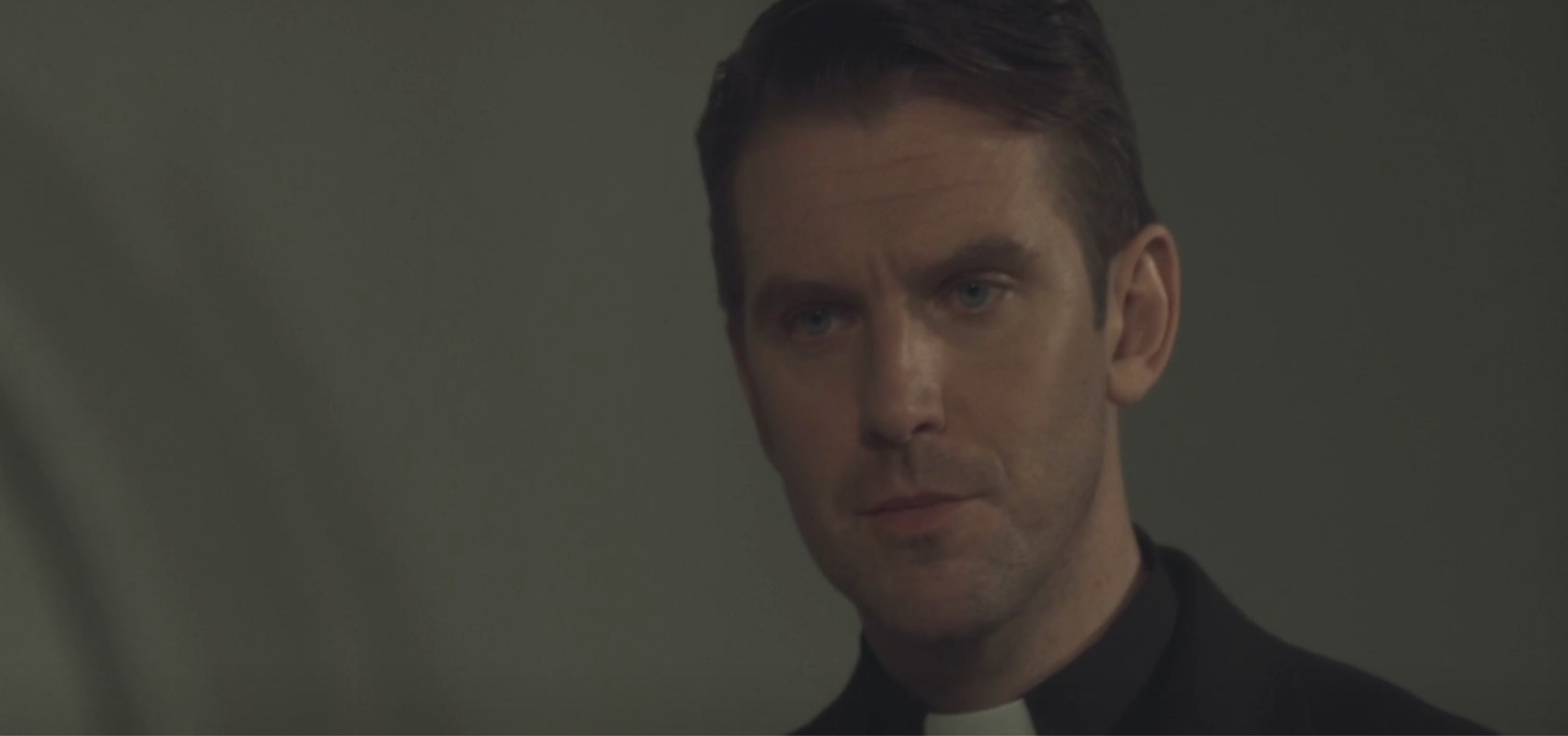 Actor portraying a somber priest looks into the distance. The image suggests a scene from a TV show or movie