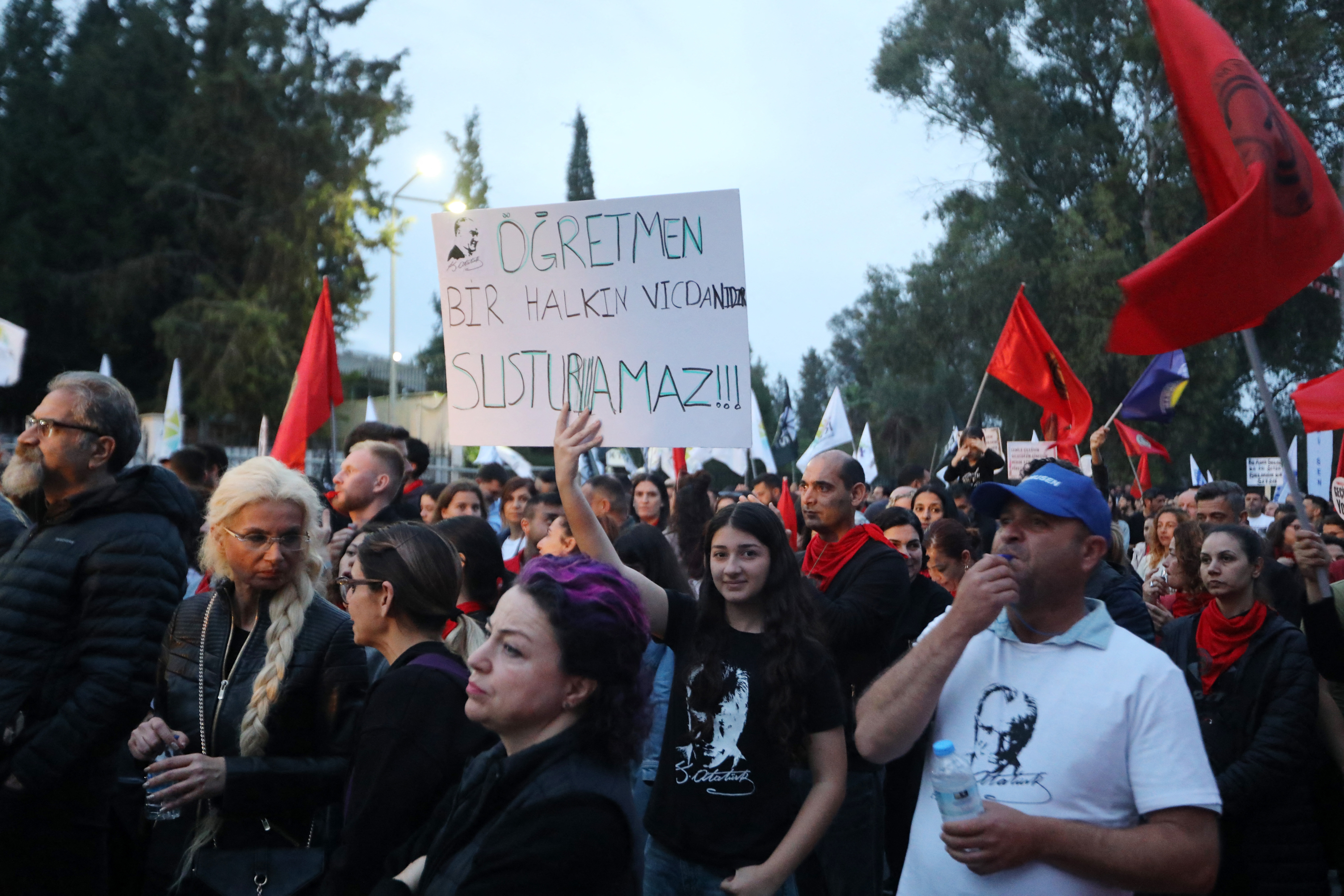 People at a protest, one holds a sign in Turkish. Some carry flags, and many are dressed casually