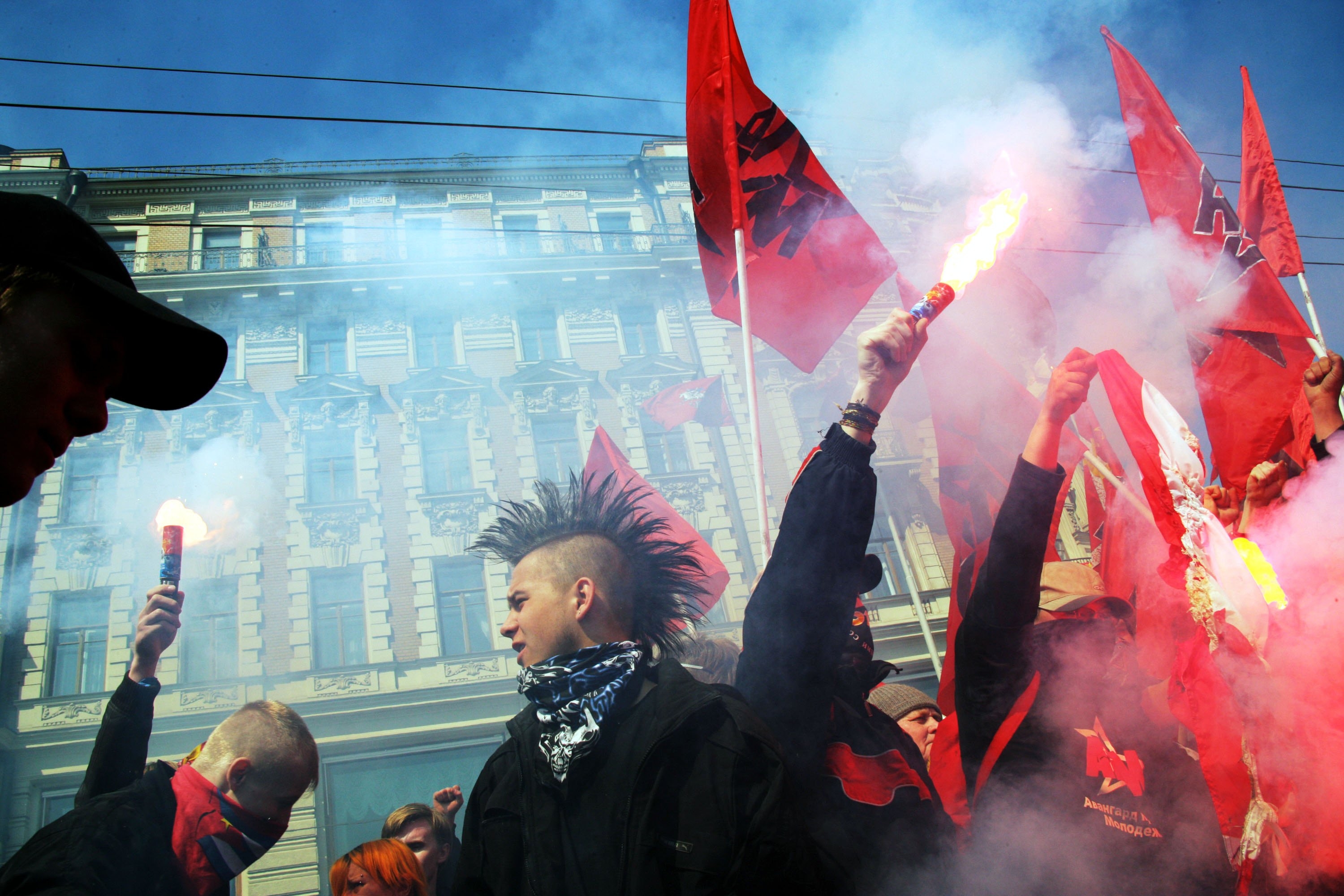 People with mohawks and bandanas holding red flags and flares in a protest or demonstration, surrounded by smoke in an urban setting