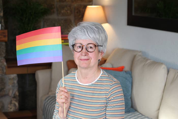 A person with short gray hair and glasses sits on a couch, smiling while holding a small rainbow flag, symbolizing LGBTQ+ pride