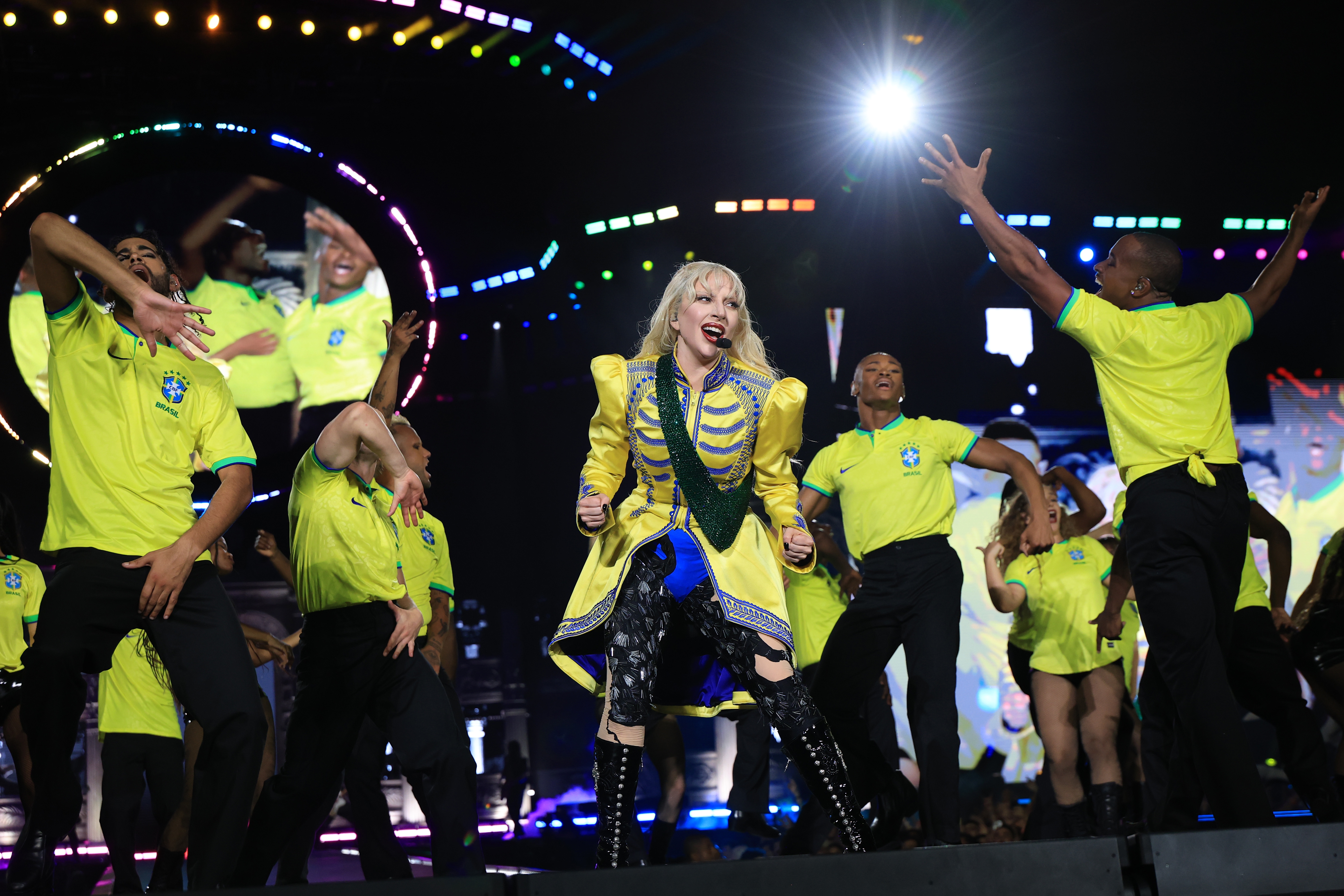 Performer in bold costume sings energetically on stage with dancers wearing matching sports shirts, set against a vibrant backdrop