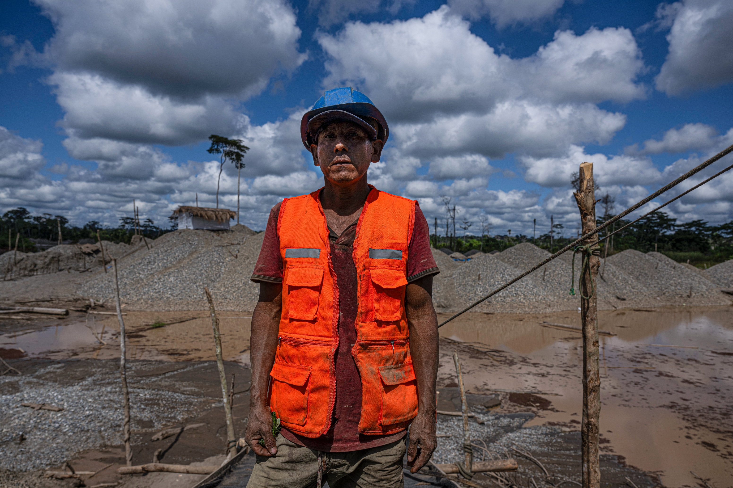 A worker wearing a hard hat and safety vest on a construction site with gravel piles and muddy ground