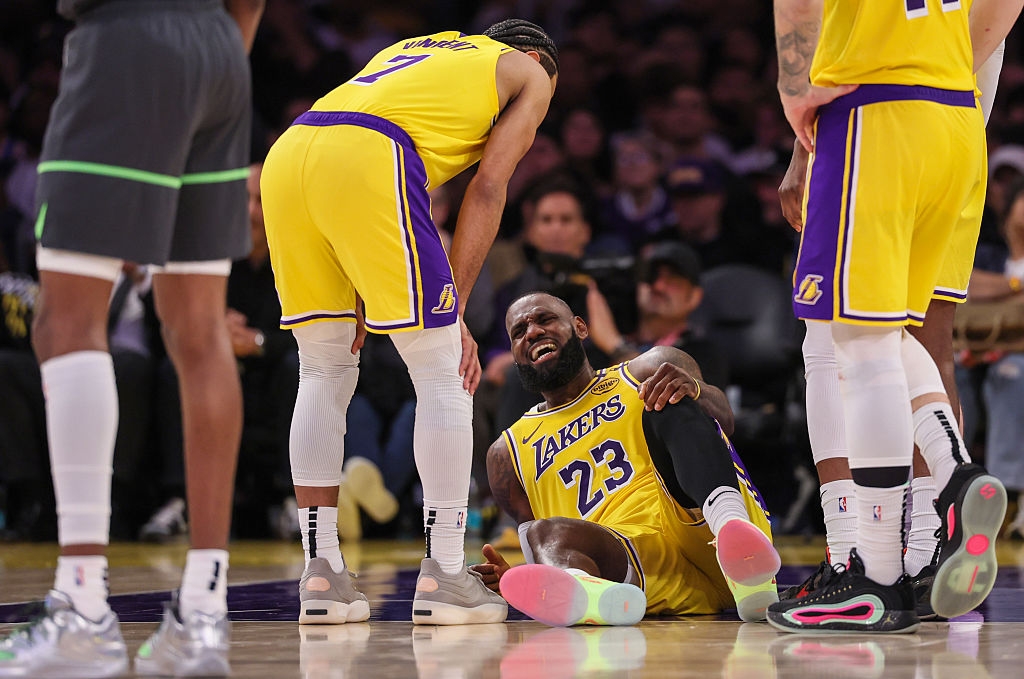 Basketball player on court, sitting and holding leg, surrounded by teammates in uniform, showing concern