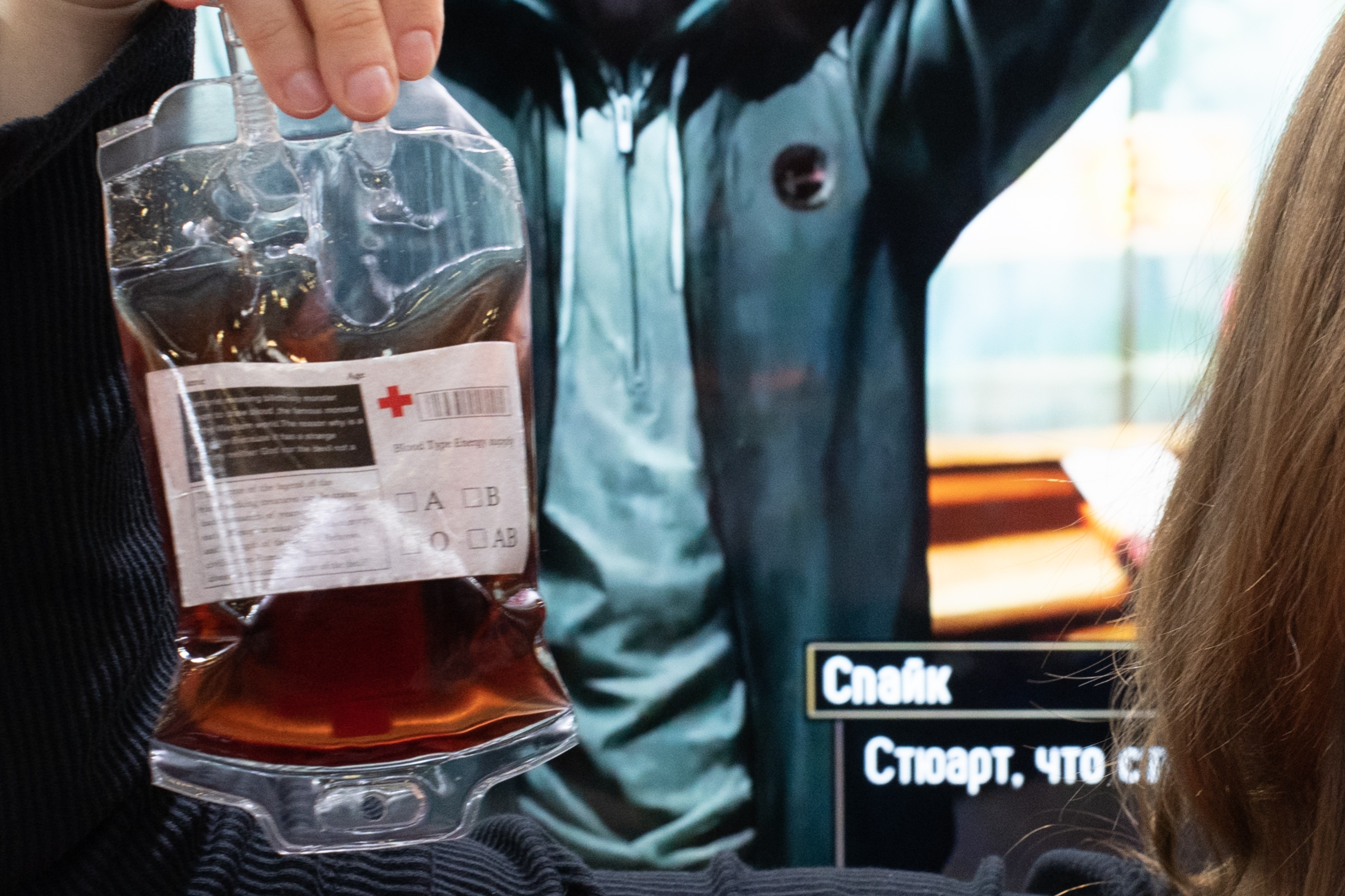 A woman holds a blood bag prop in front of a video game display featuring a character with a hood