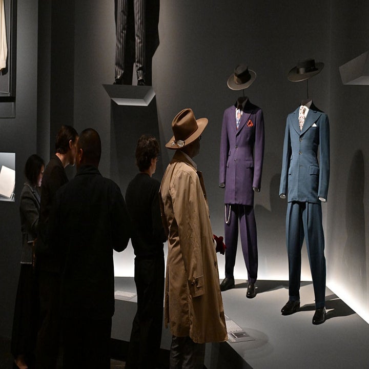 People examine two mannequins displaying vintage suits with hats. The setting appears to be an exhibition or museum