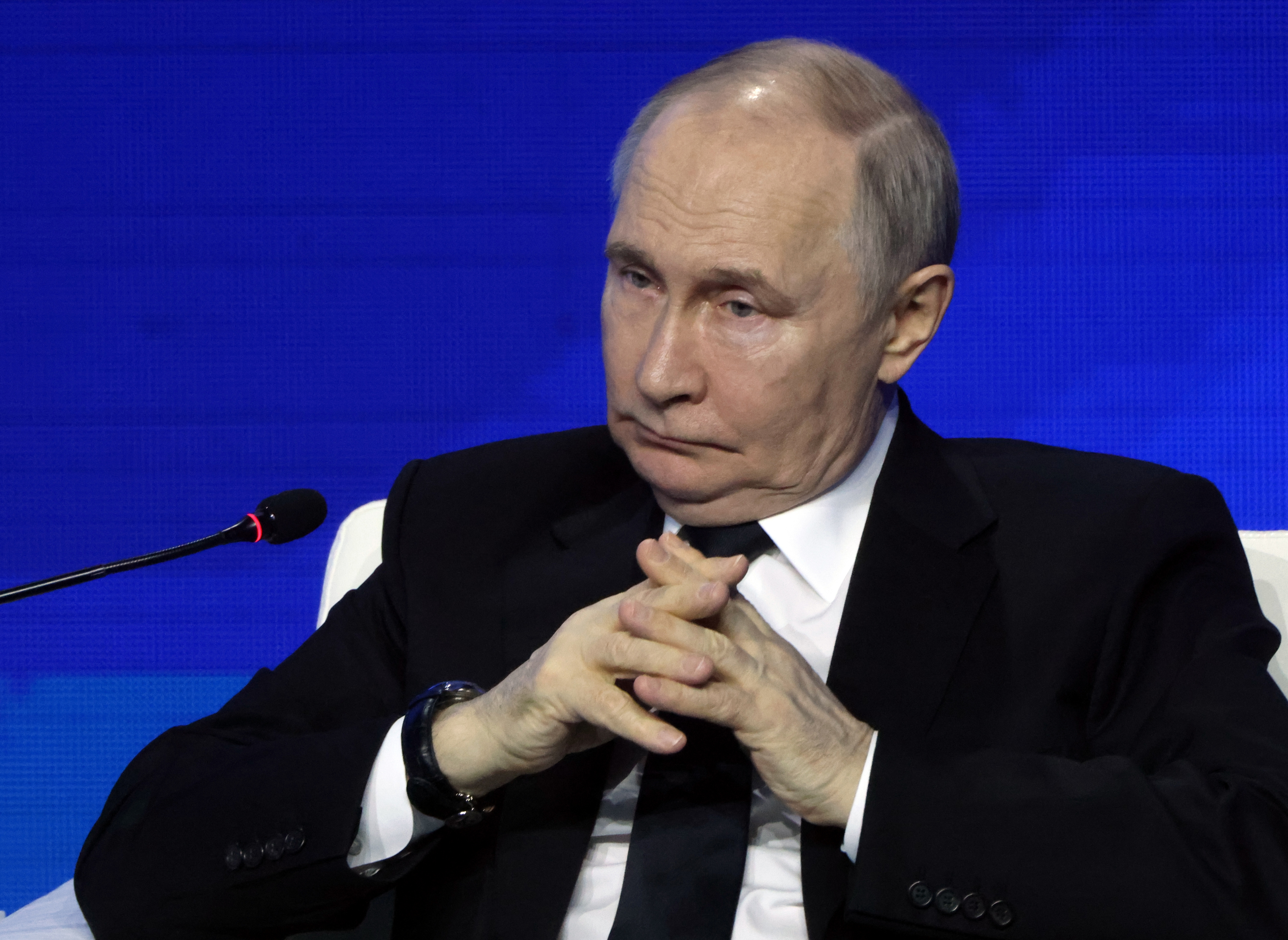 A person in a suit sits with hands clasped, appearing contemplative, in front of a microphone