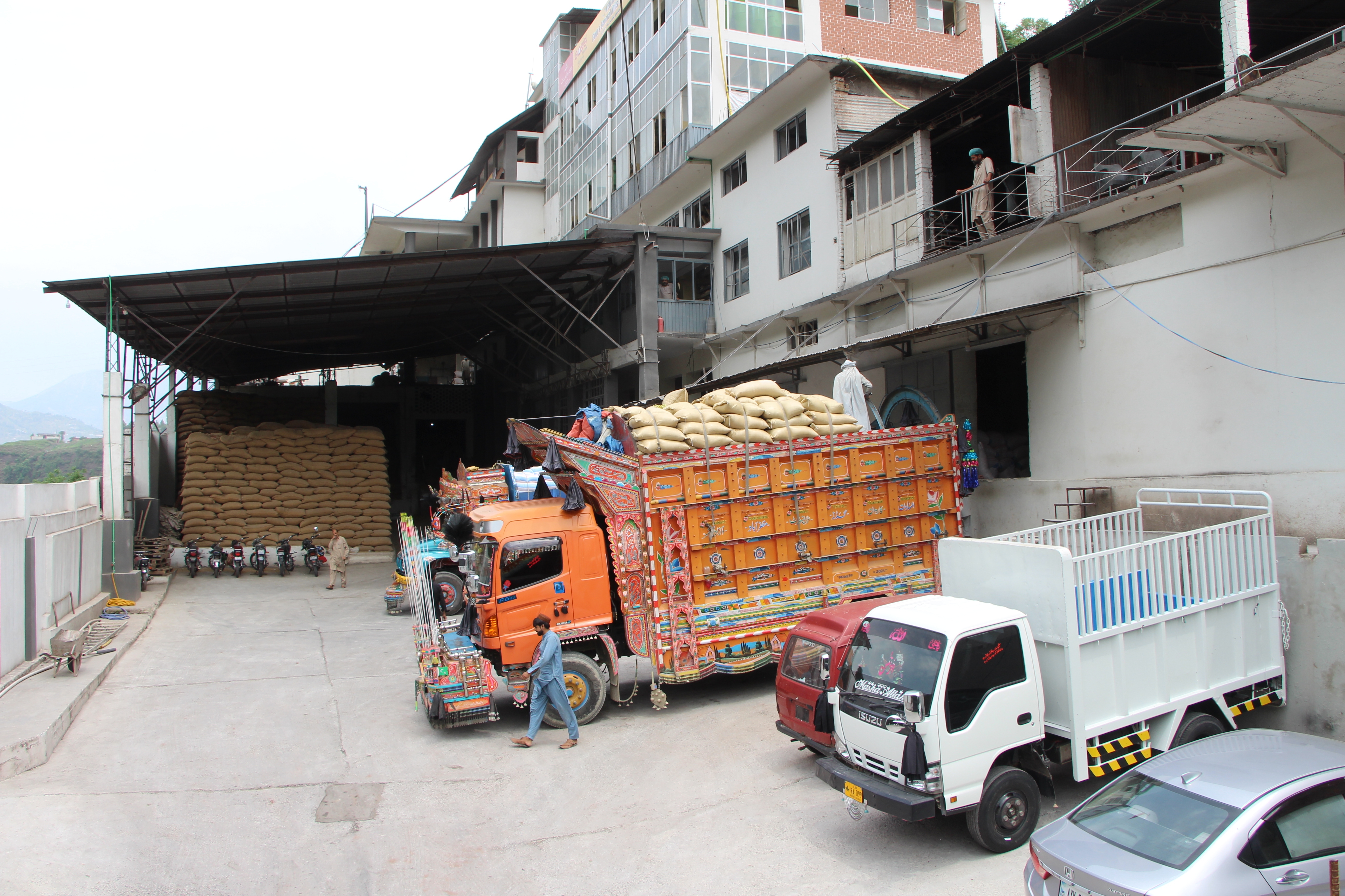 Trucks loaded with stacked bags are parked near a warehouse, with workers visible on the loading dock