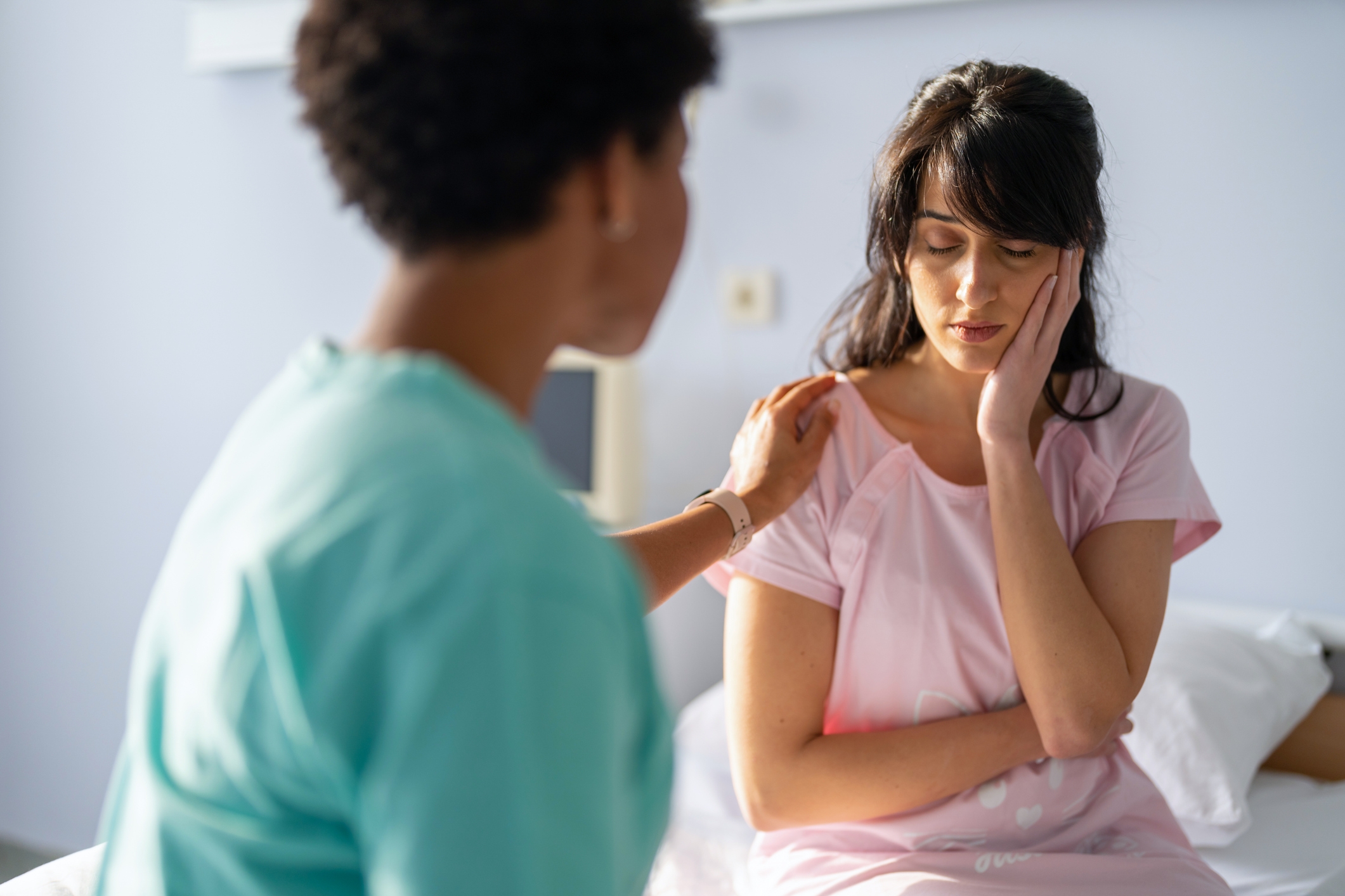 A person in a medical uniform comforts a woman sitting with her head resting on her hand, looking concerned