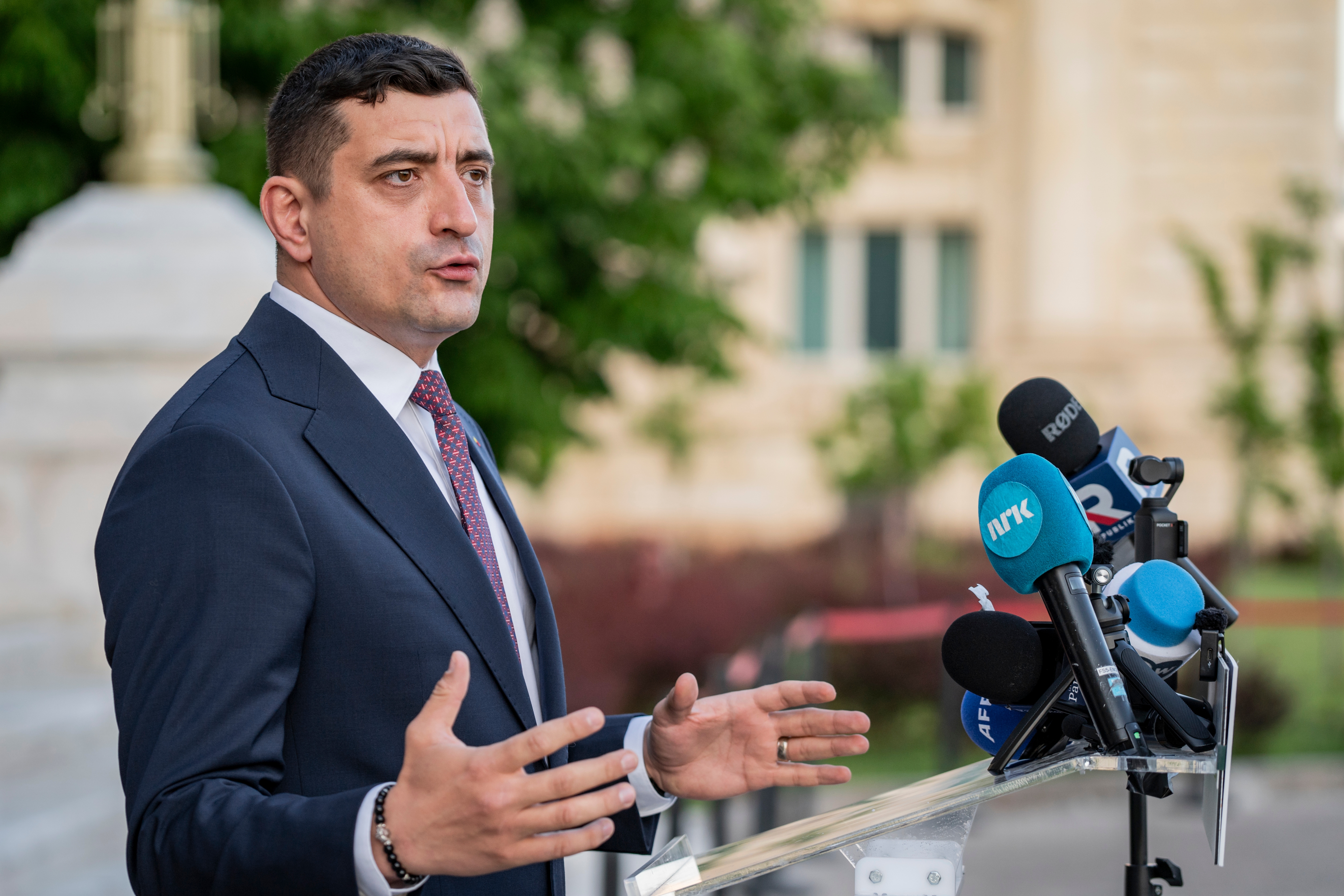 A person in a suit speaks at a podium with multiple microphones outside a building