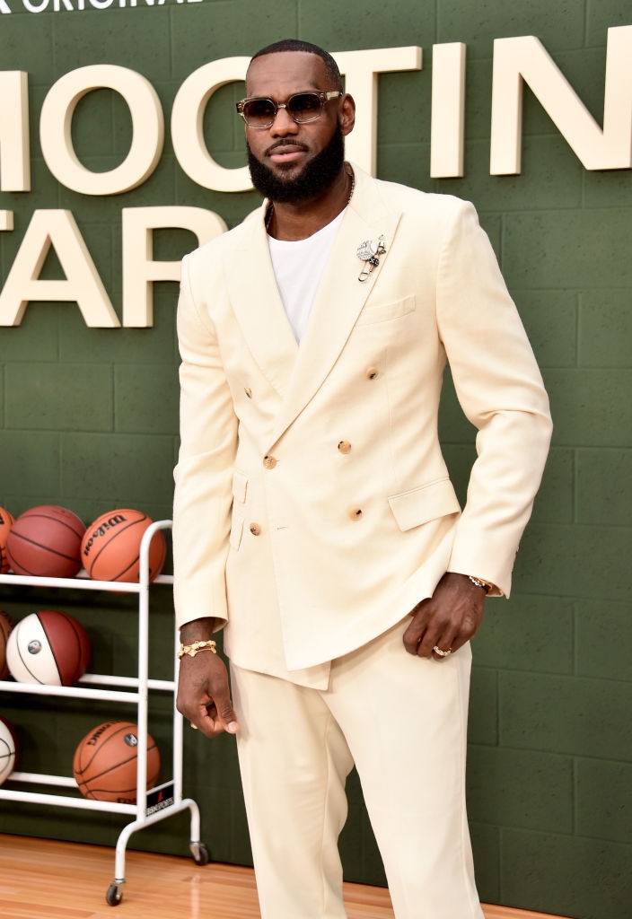 Person in a double-breasted suit with sunglasses stands confidently at an event, with basketballs in the background