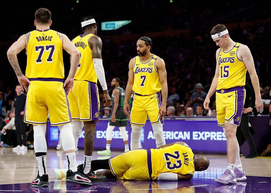 Basketball player lies on the court while teammates in jerseys gather around, showing concern during a game