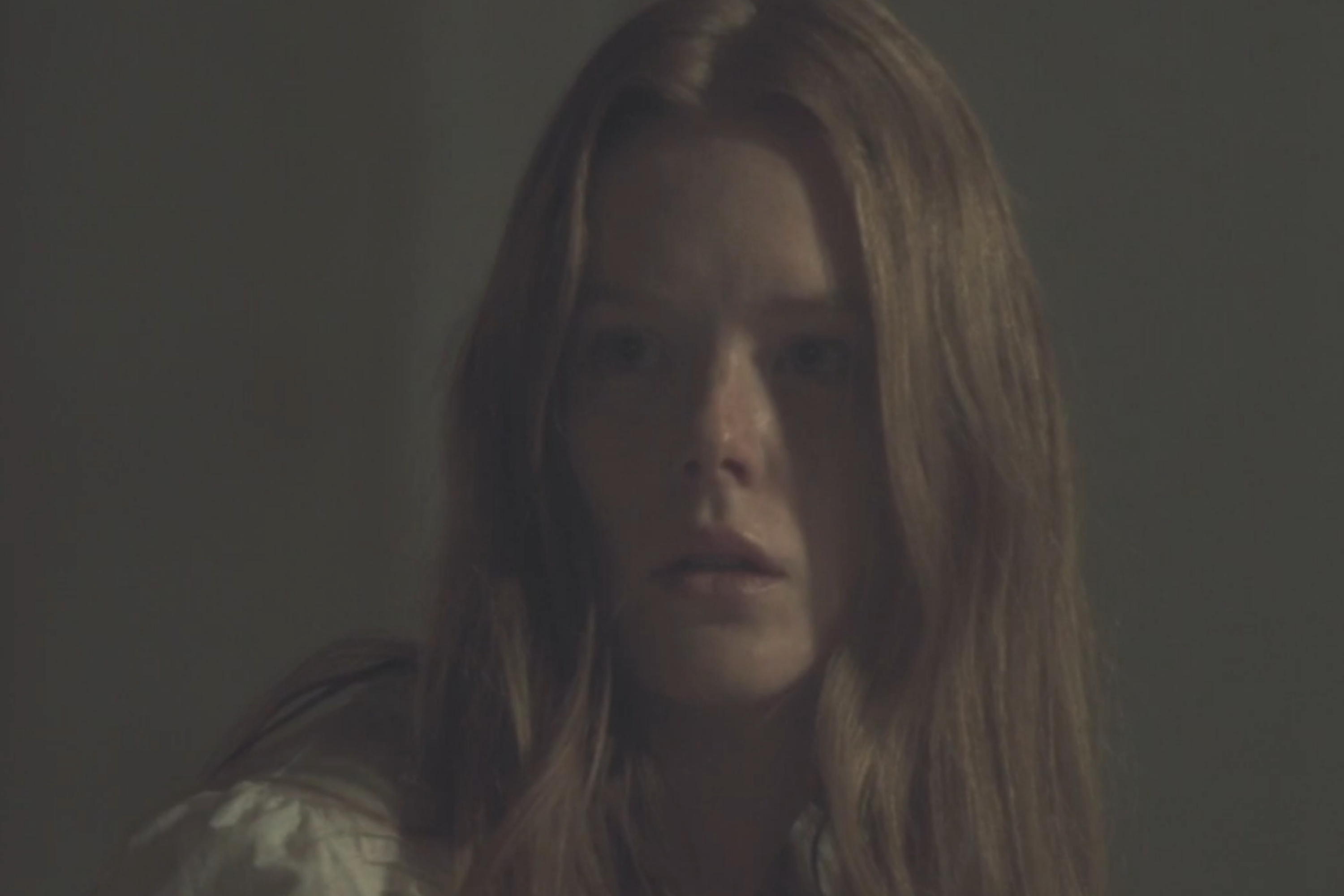 A person with long hair looks concerned in a dimly lit room, wearing a casual top