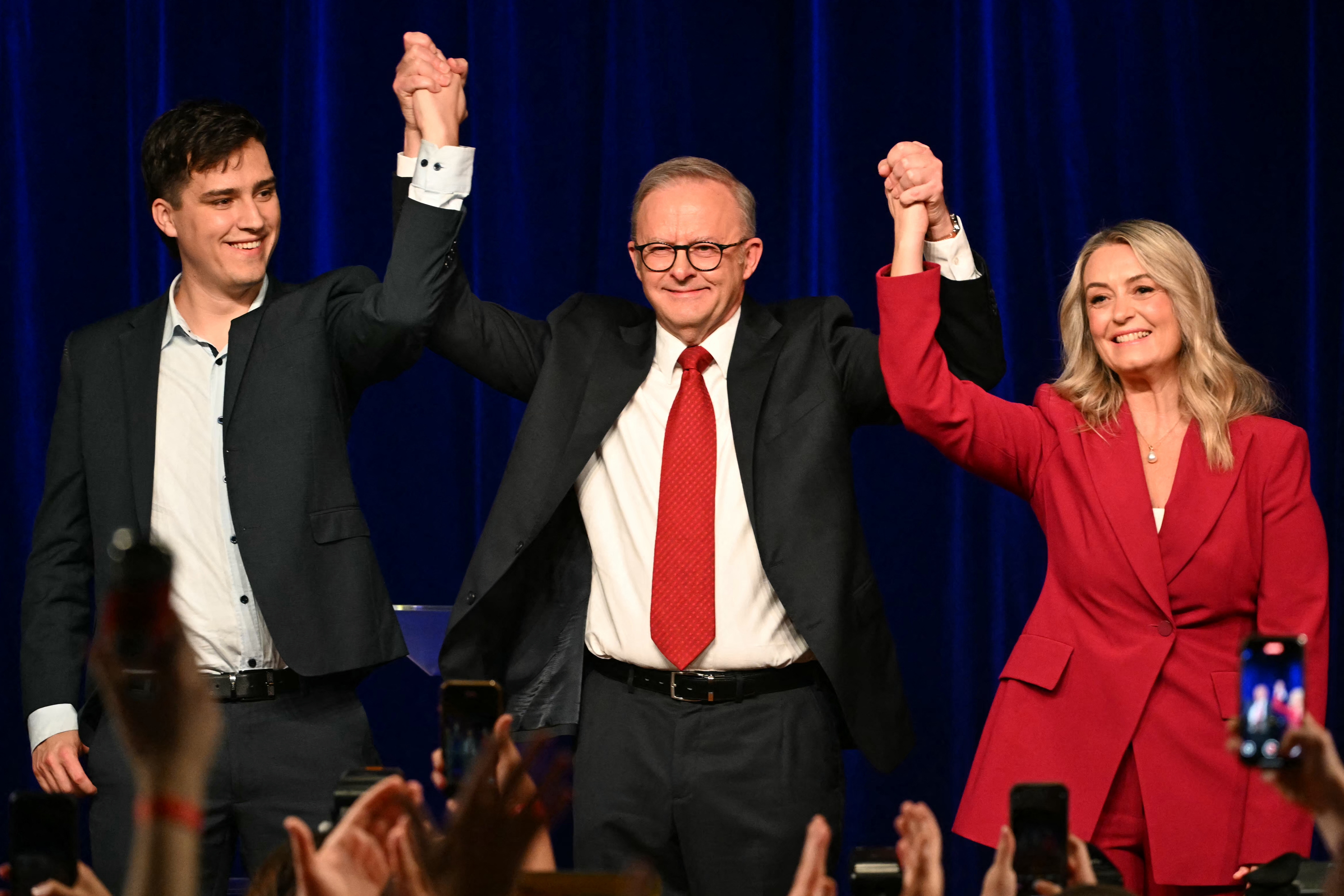 Three people on stage celebrate, holding hands up. The center person wears a suit, the person on the right wears a red suit. Audience holds phones up