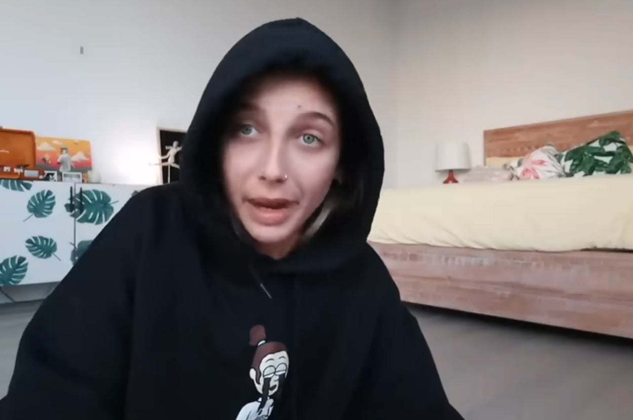 Person in a hoodie sitting in a bedroom with a bed and dresser in the background
