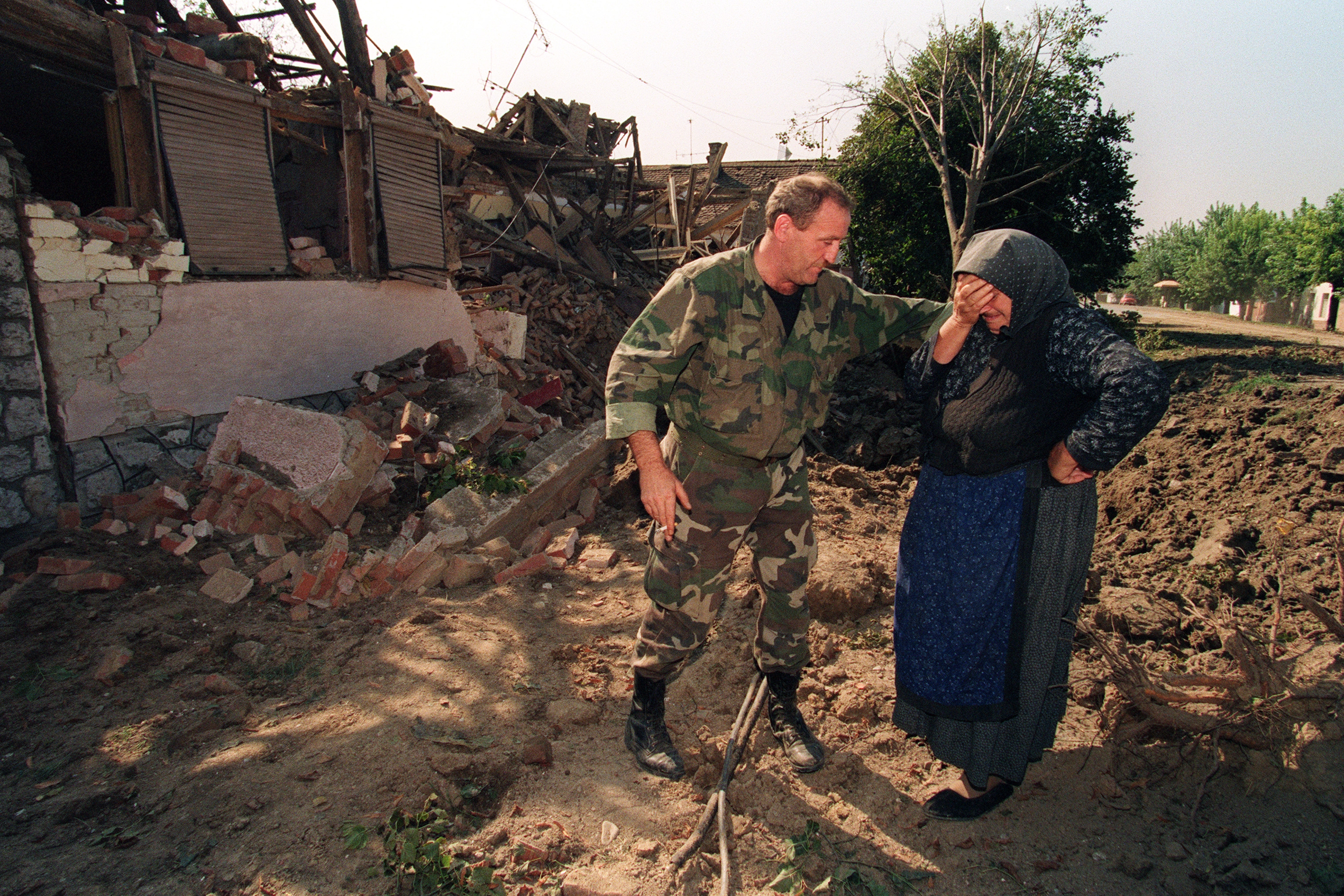 A soldier consoles a distressed woman near rubble of a destroyed building, highlighting aftermath of destruction and human impact