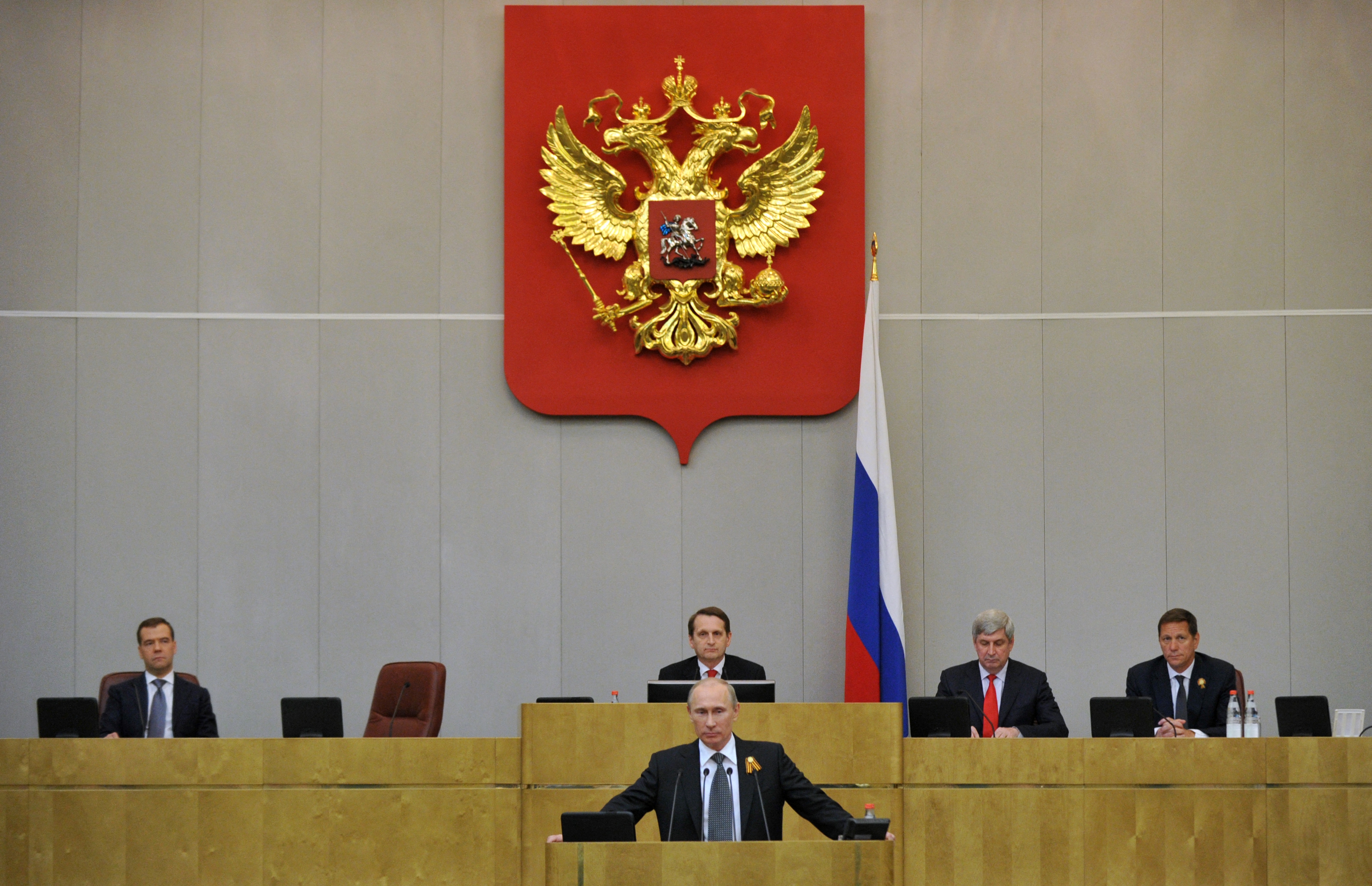 A speaker addresses an assembly in a formal government chamber, featuring the Russian coat of arms and seated officials at a podium