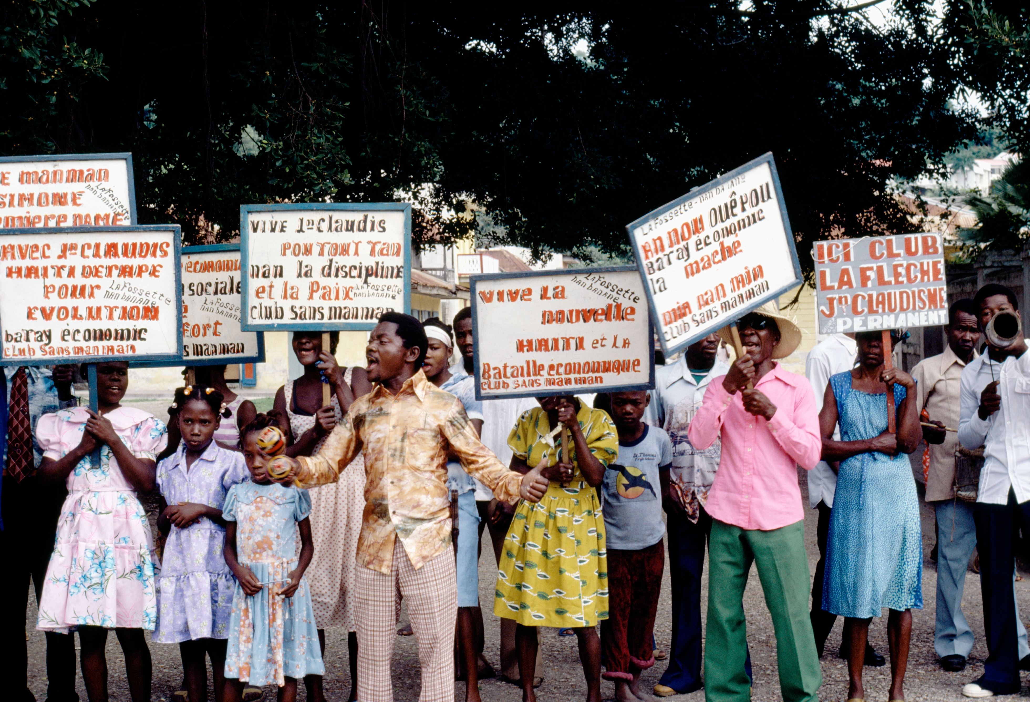 A group of people holding various protest signs in a public area, including adults and children, some playing musical instruments
