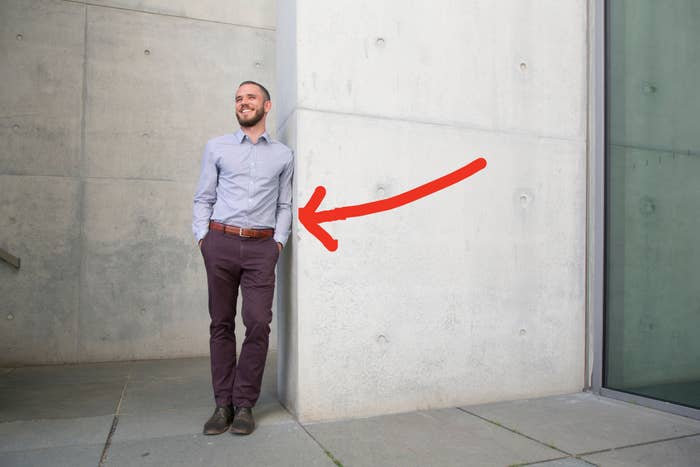A person in business casual attire is leaning against a concrete wall, looking off into the distance with a smile