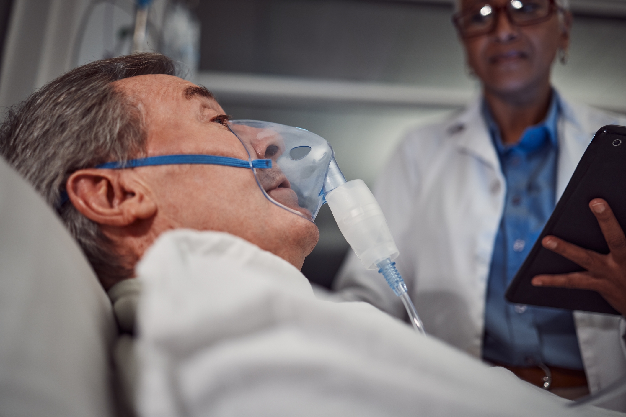 A patient wearing an oxygen mask lies in a hospital bed, while a doctor holds a tablet nearby