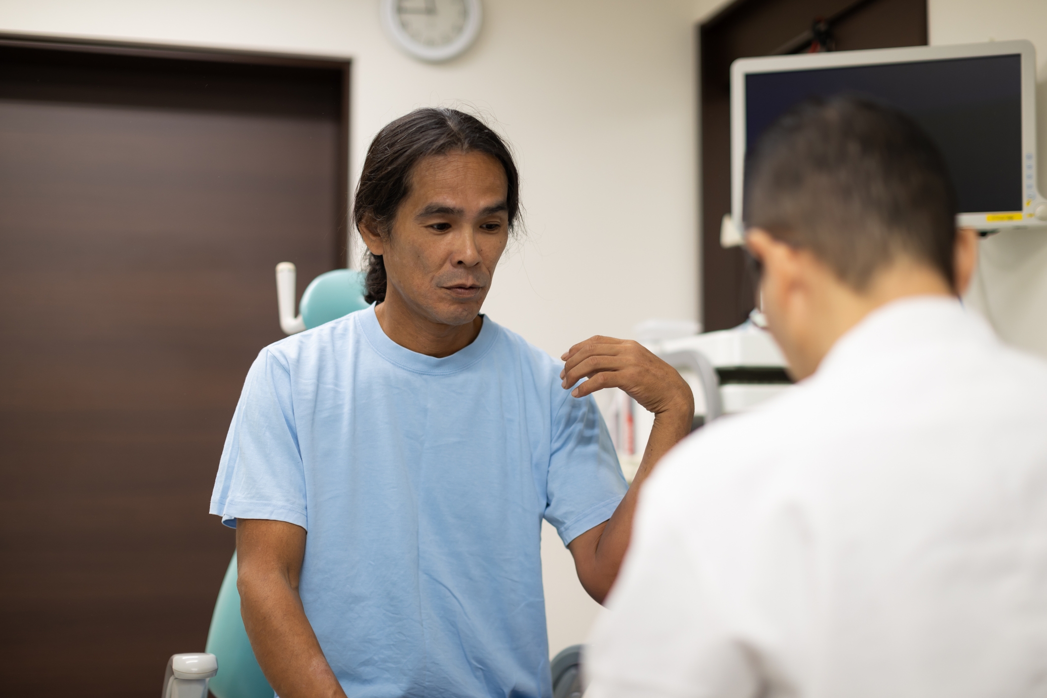 A man in a blue shirt gestures while talking to another man in an office setting, with a clock and computer in the background