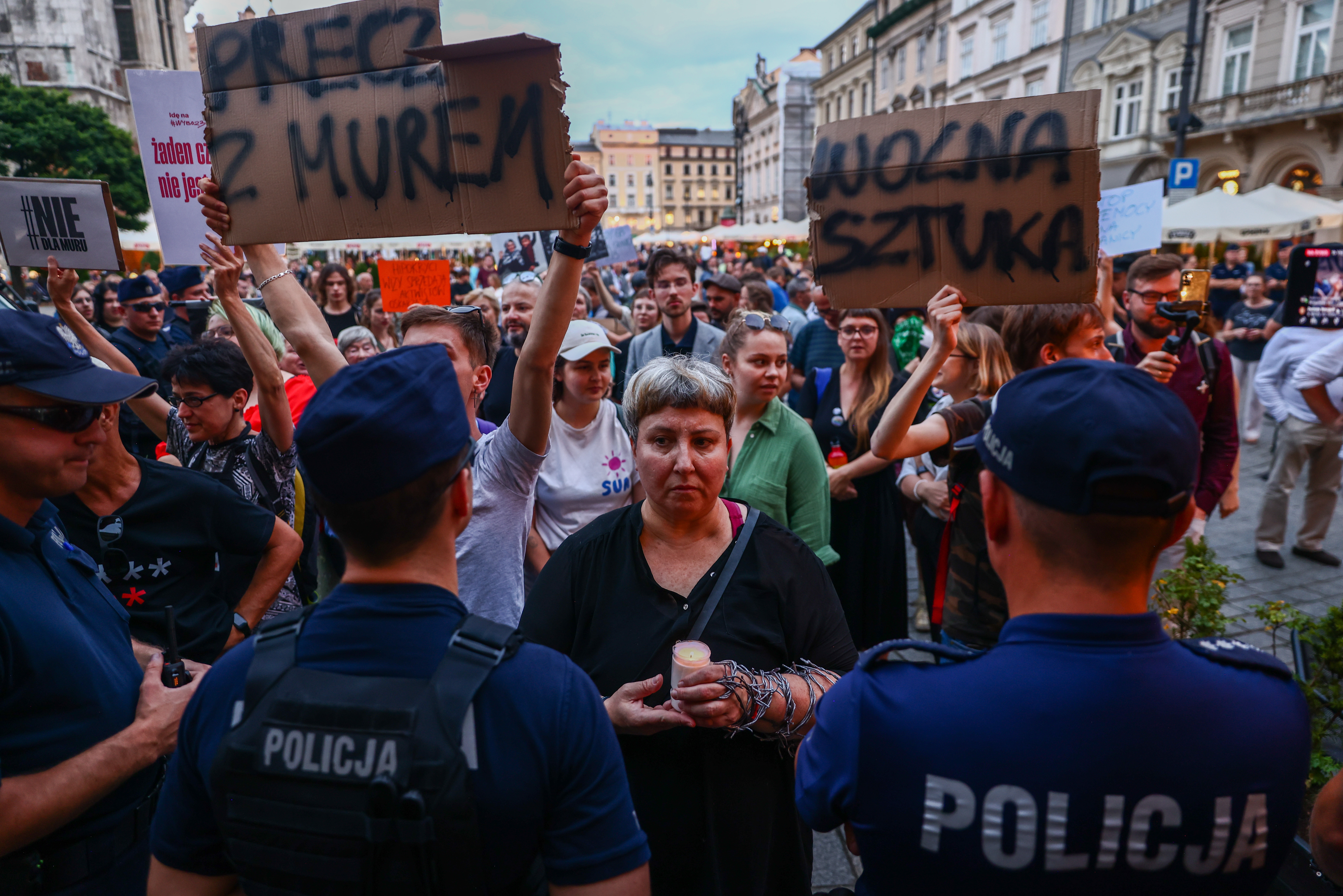 Protesters holding signs face police officers in a crowded street demonstration. A person in the center holds a candle