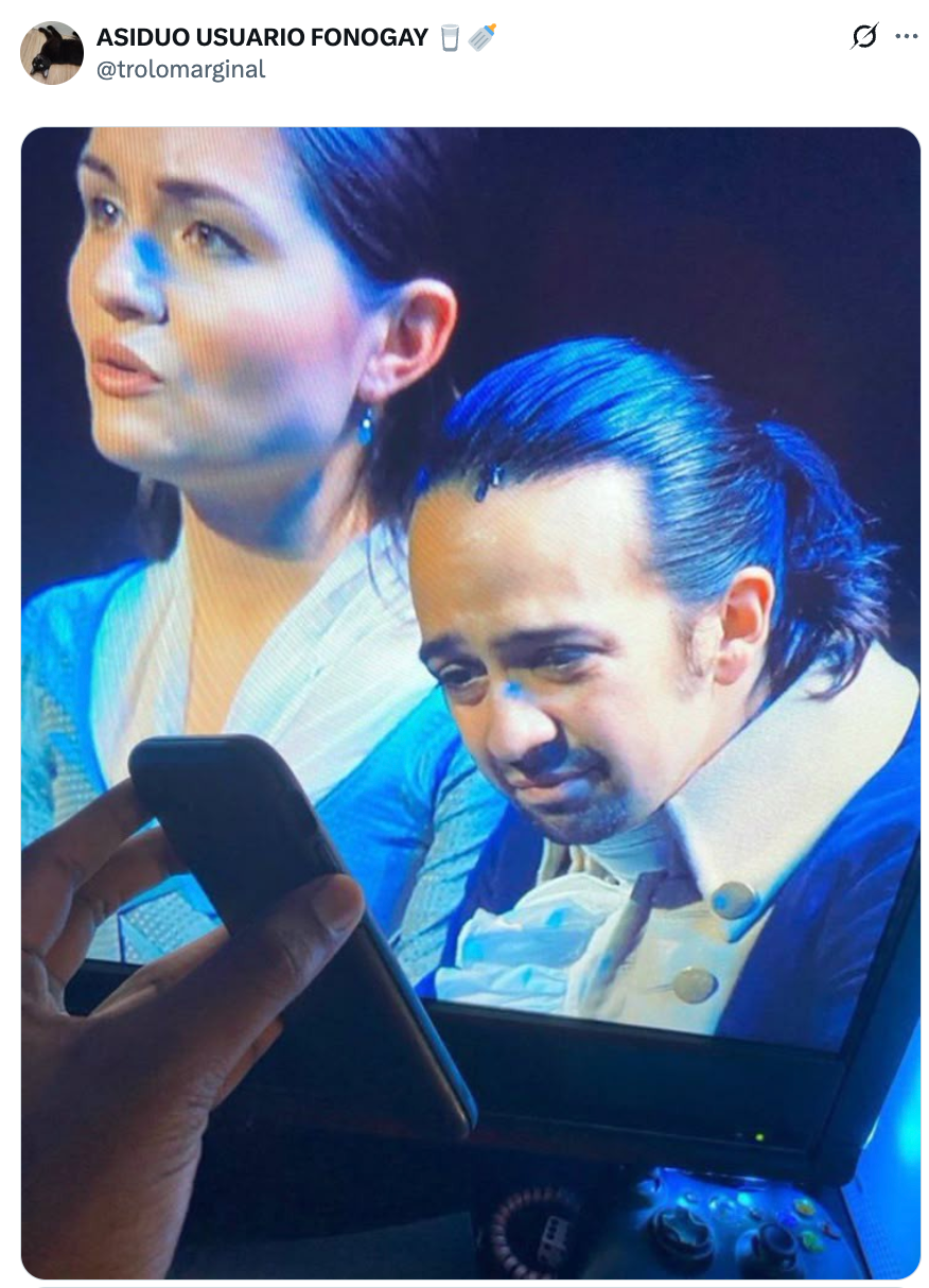 Two performers in period costumes sing passionately on stage, with a hand holding a mobile phone in the foreground