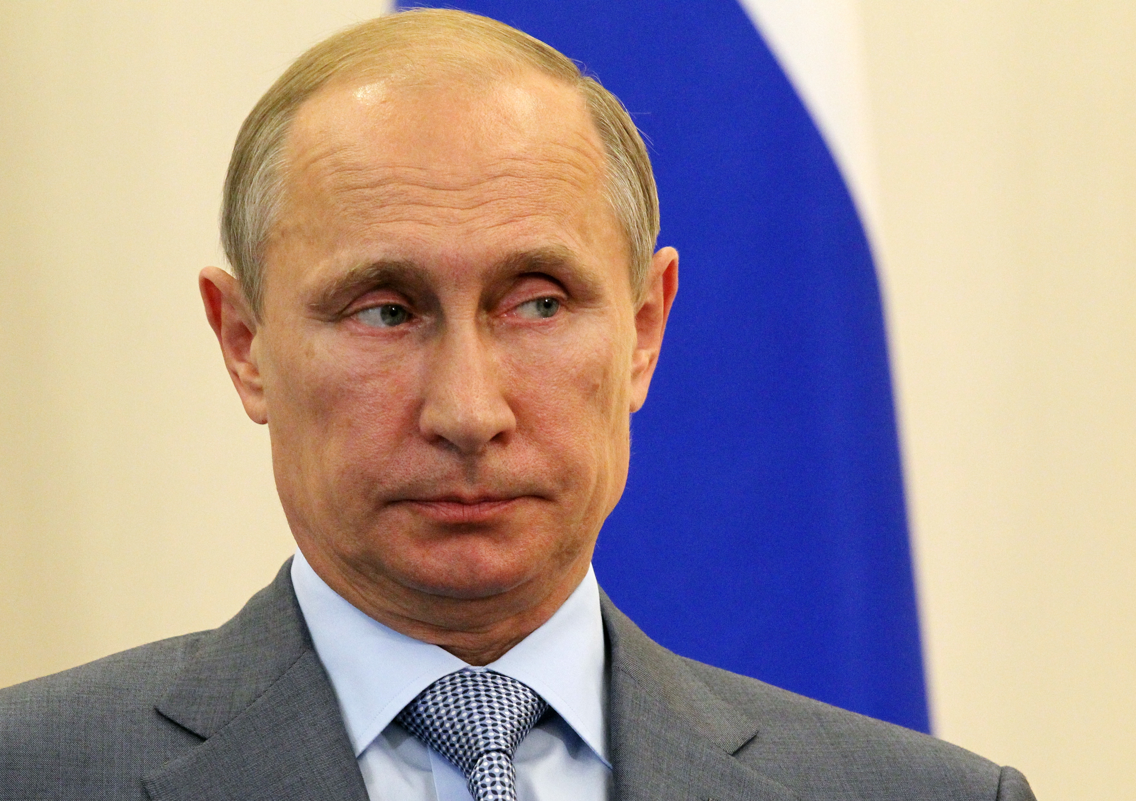 Man in a suit looking slightly to the side, with a neutral expression. A patterned background is partially visible