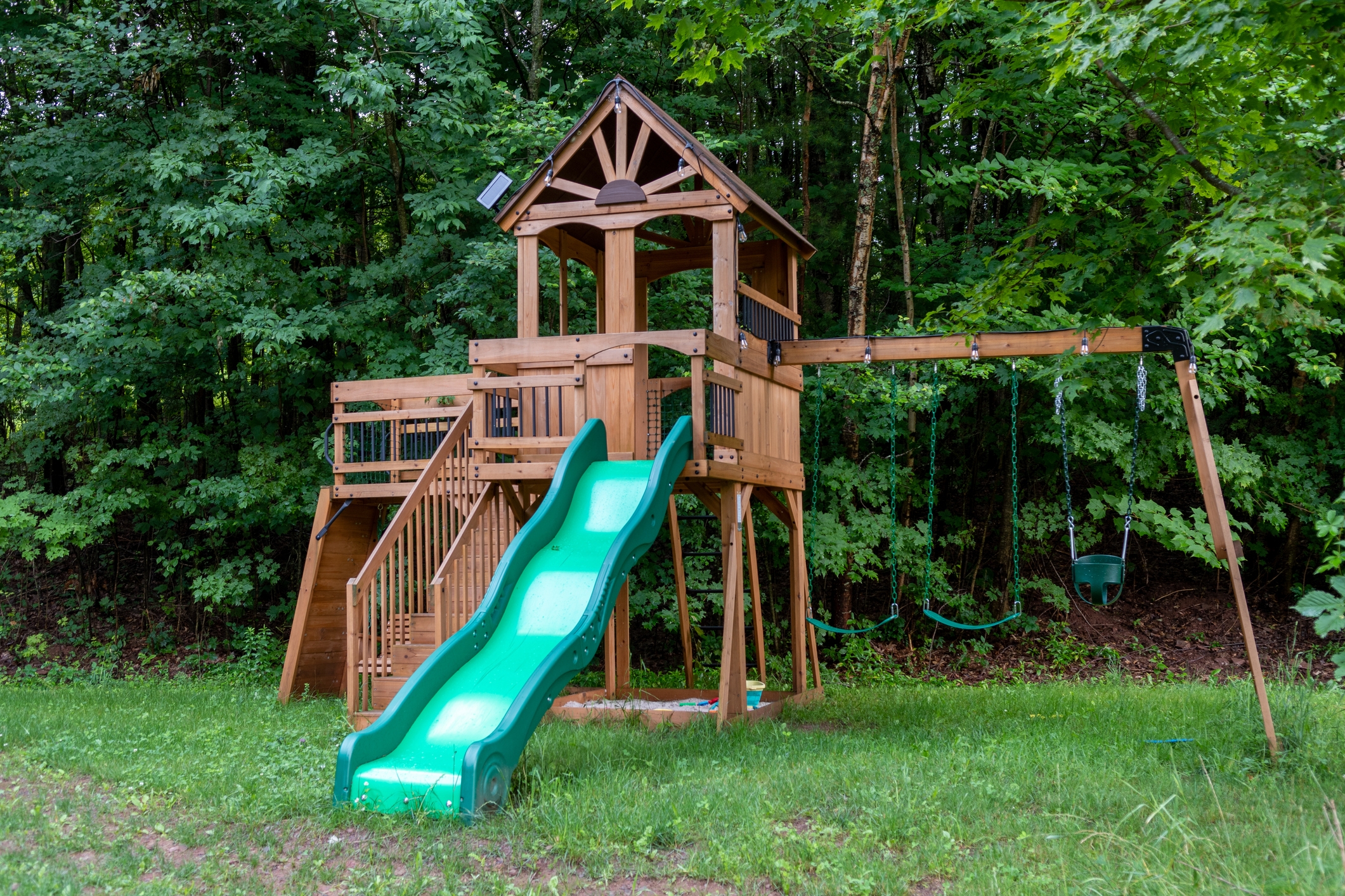 Wooden playset with a slide, swings, and a small tower, surrounded by lush green trees in the background