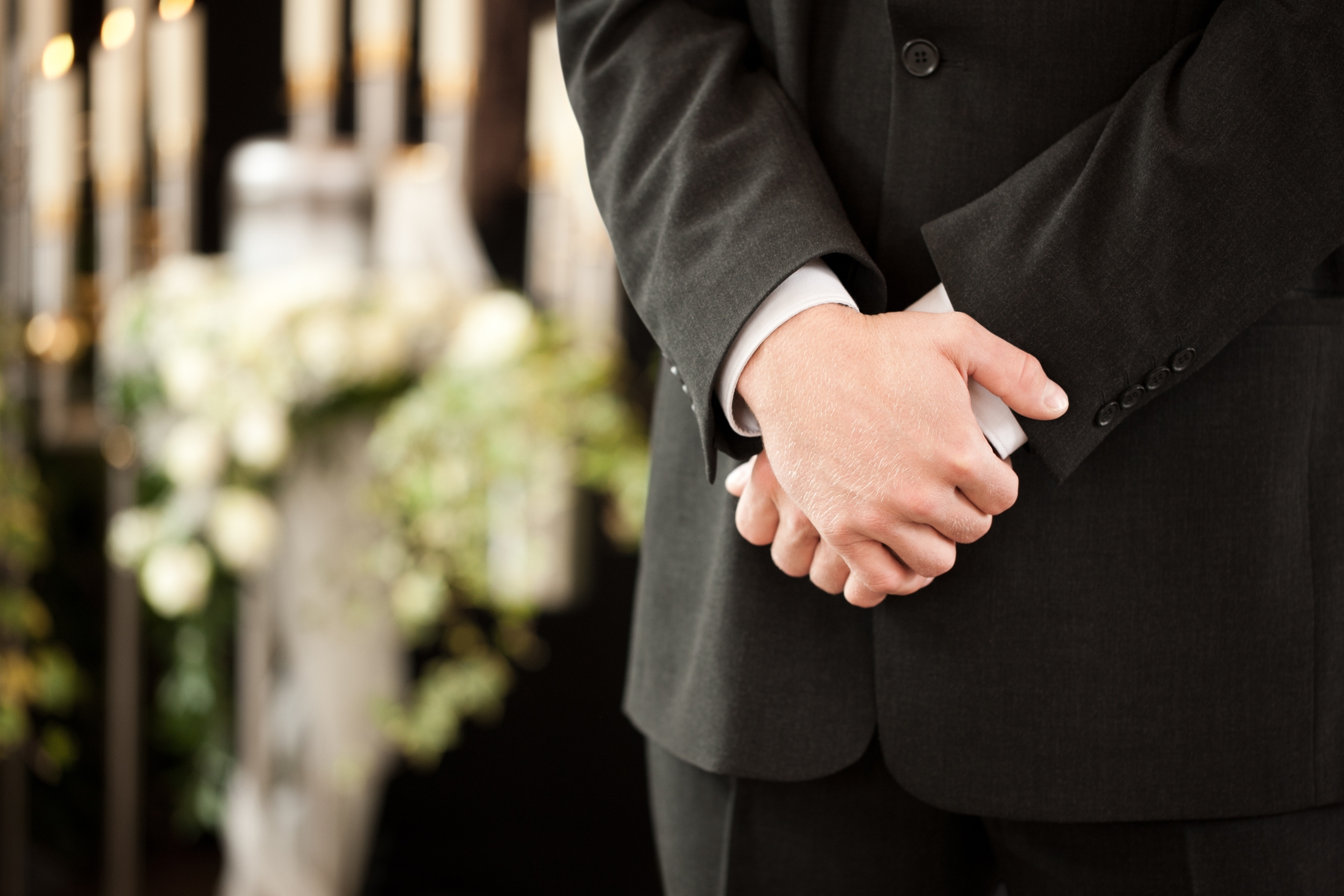Person in formal suit standing with hands clasped, blurred floral arrangement in the background
