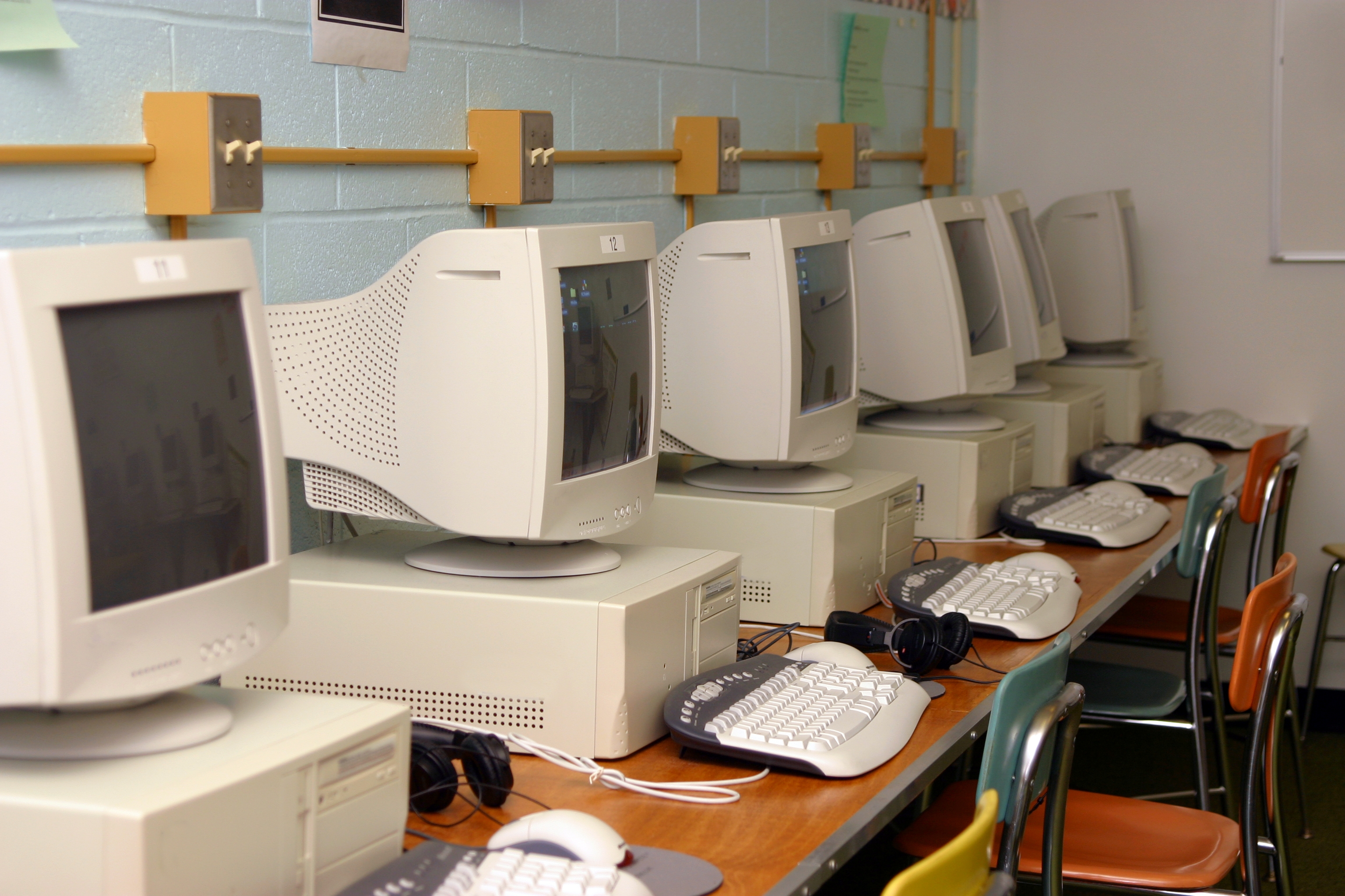 Row of vintage CRT monitors and computers lined up in a classroom setting, with keyboards, mice, and headphones on the desk