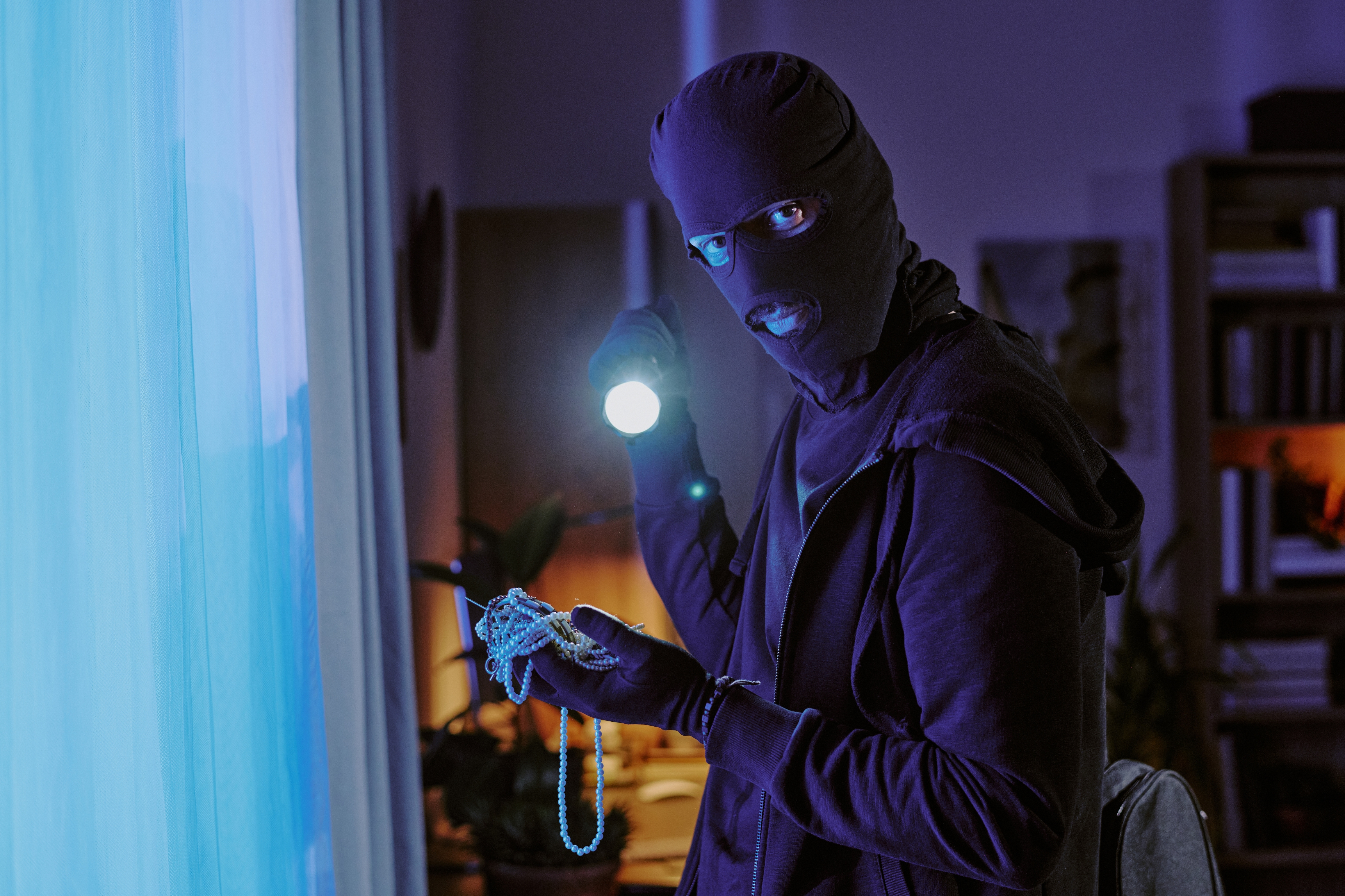 Person in a dark outfit and mask holds a flashlight and pearl necklace, appearing to be a burglar. The room is dimly lit with bookshelves in the background
