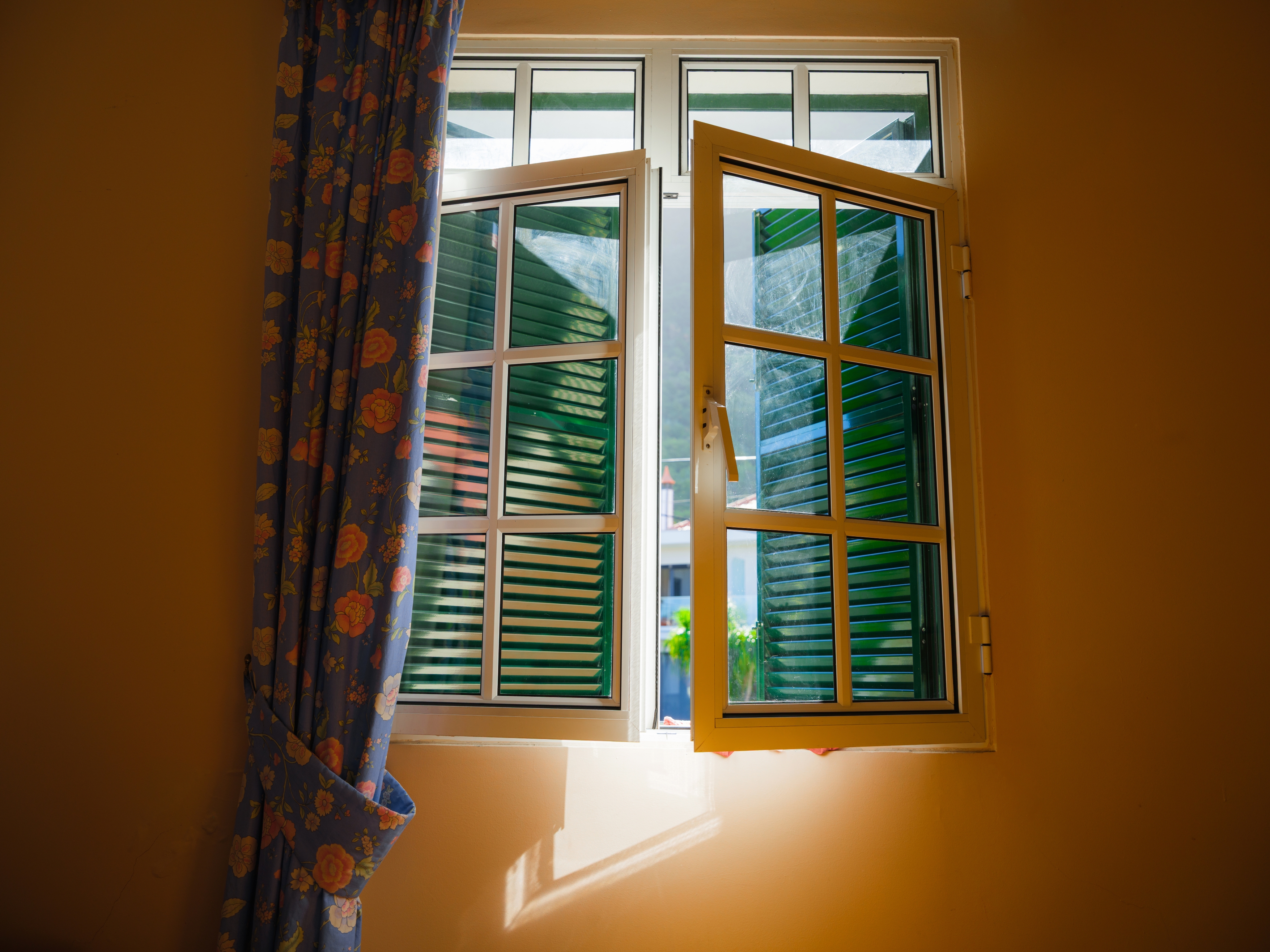A partially open window with green shutters allows sunlight to pour into a room with floral-patterned curtains