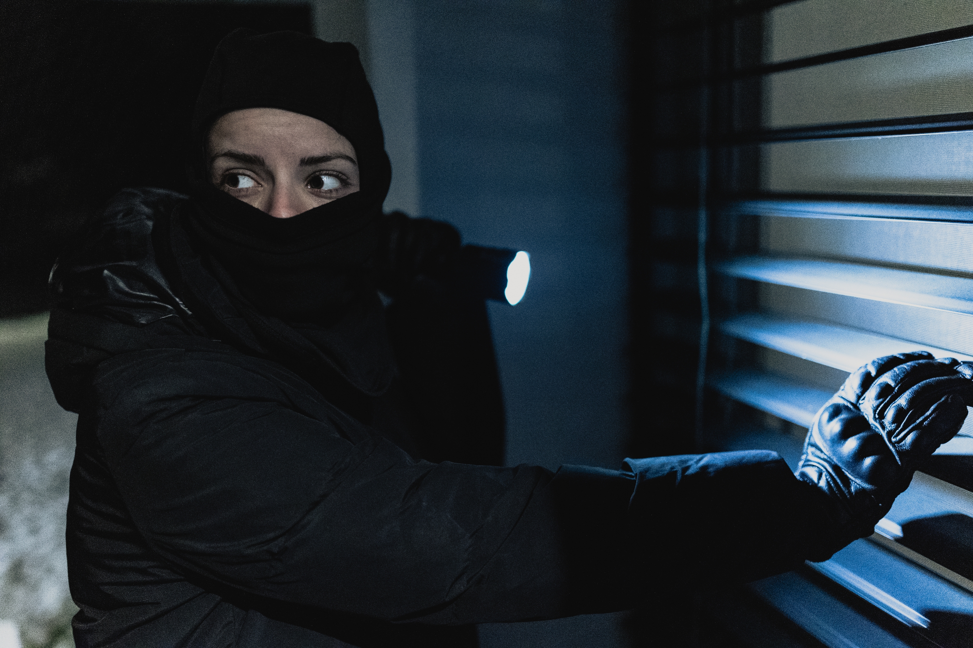 Person in dark clothing and gloves peeks through blinds while holding a flashlight, appearing cautious and focused