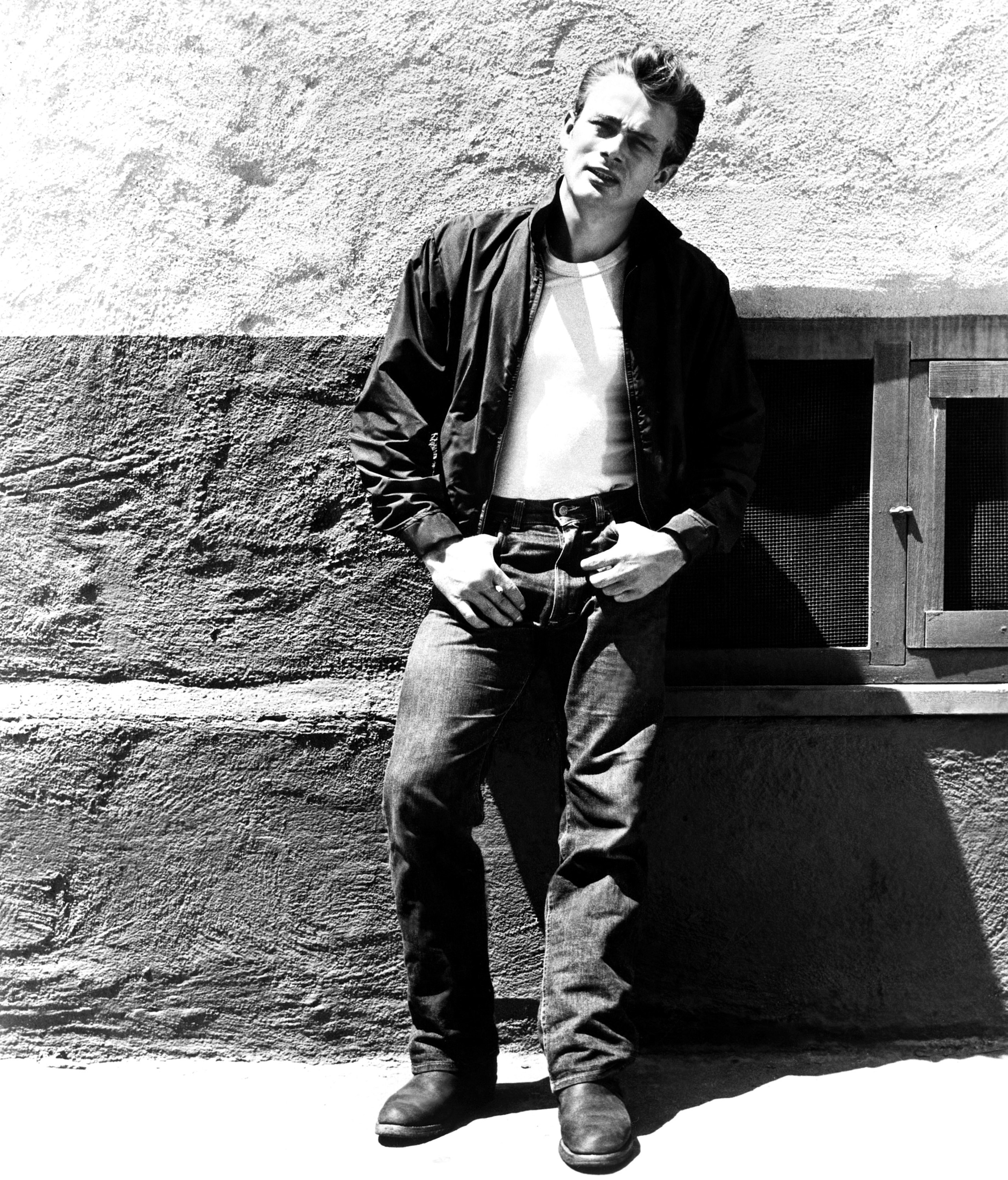 James Deen standing against a wall, wearing a leather jacket, white shirt, jeans, and boots, gazing forward with a relaxed posture