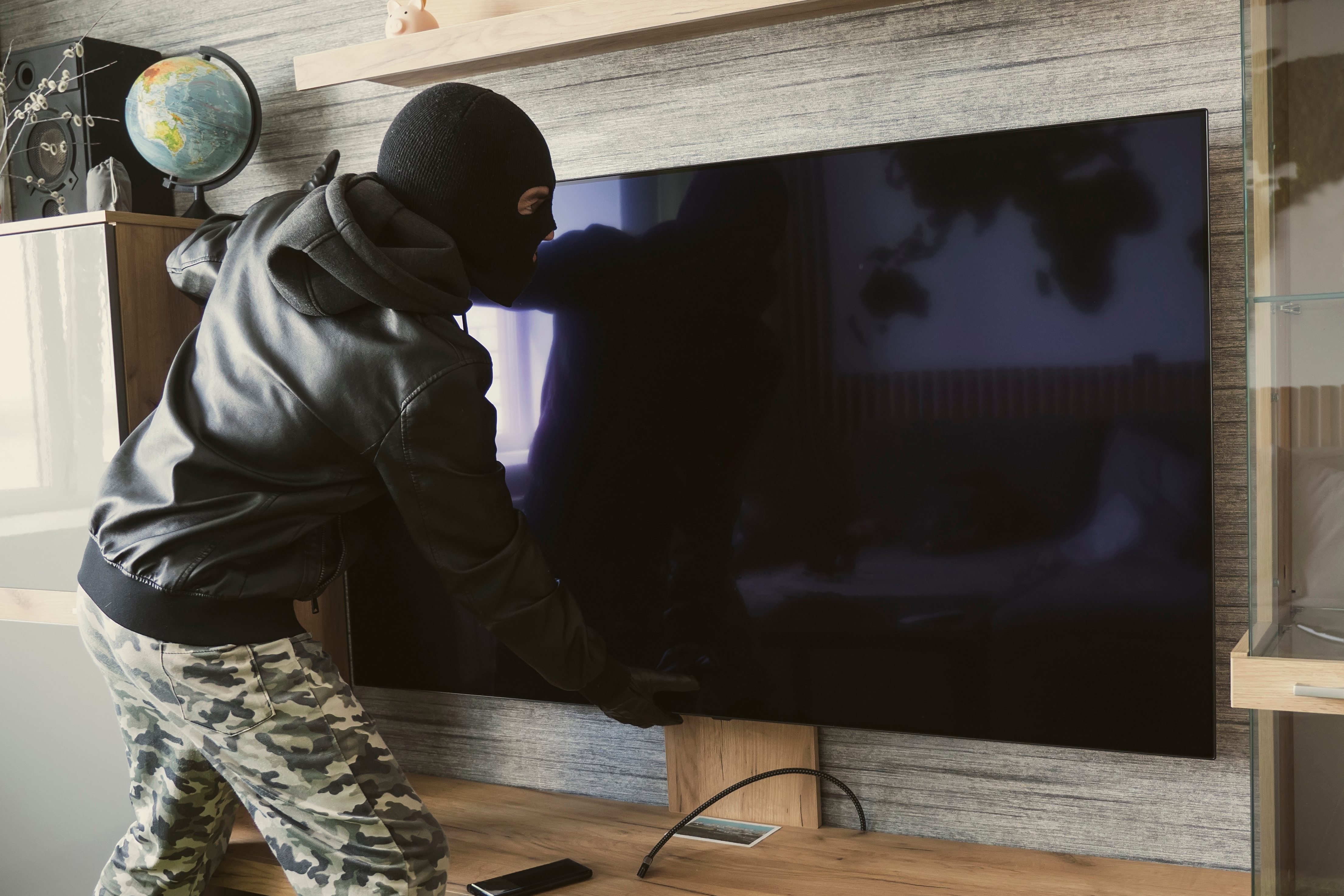 A person in dark clothing and a mask appears to be stealing a large flat-screen TV from a modern living room setting