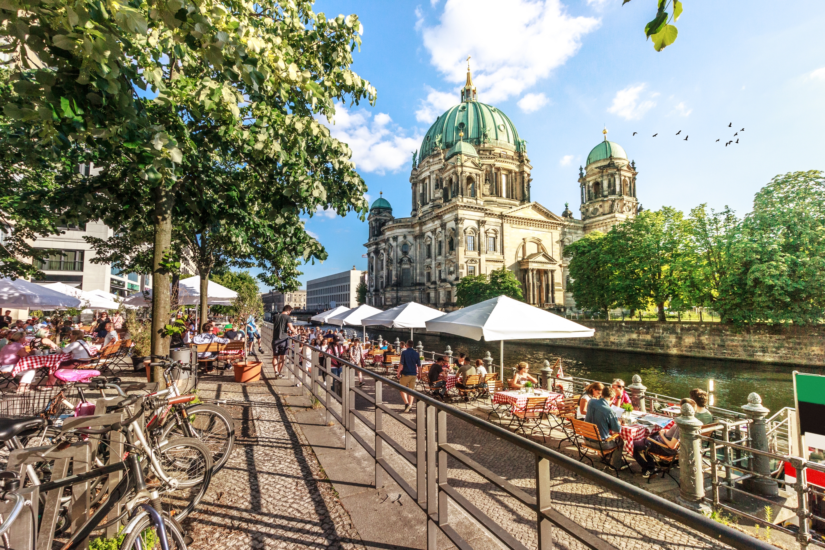 Cafe patrons sit along a riverside with a historic domed cathedral in the background, under a clear sky with greenery and bicycles nearby