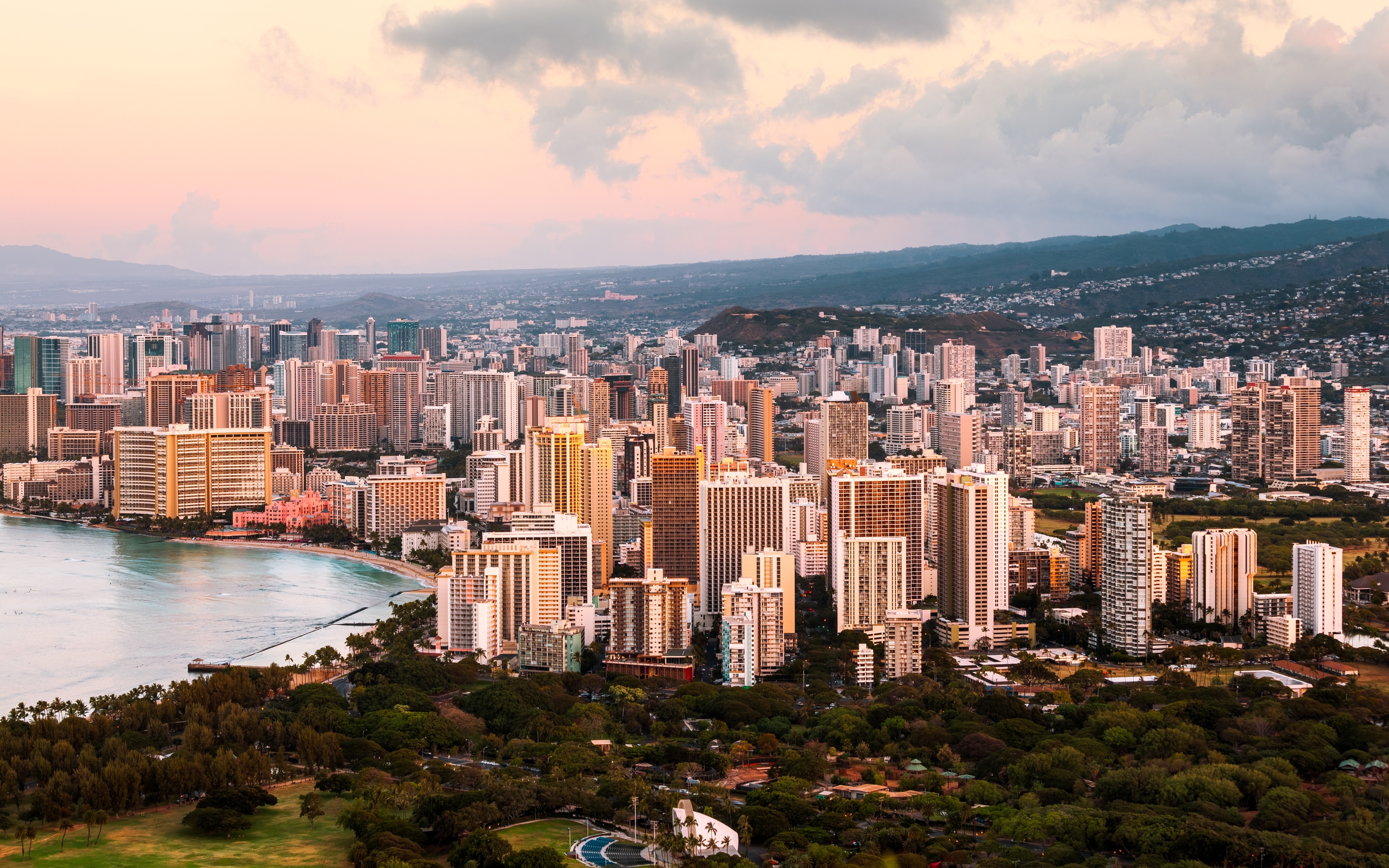 Aerial view of a bustling city skyline with numerous tall buildings along a curved coastline, surrounded by lush greenery and mountains in the background