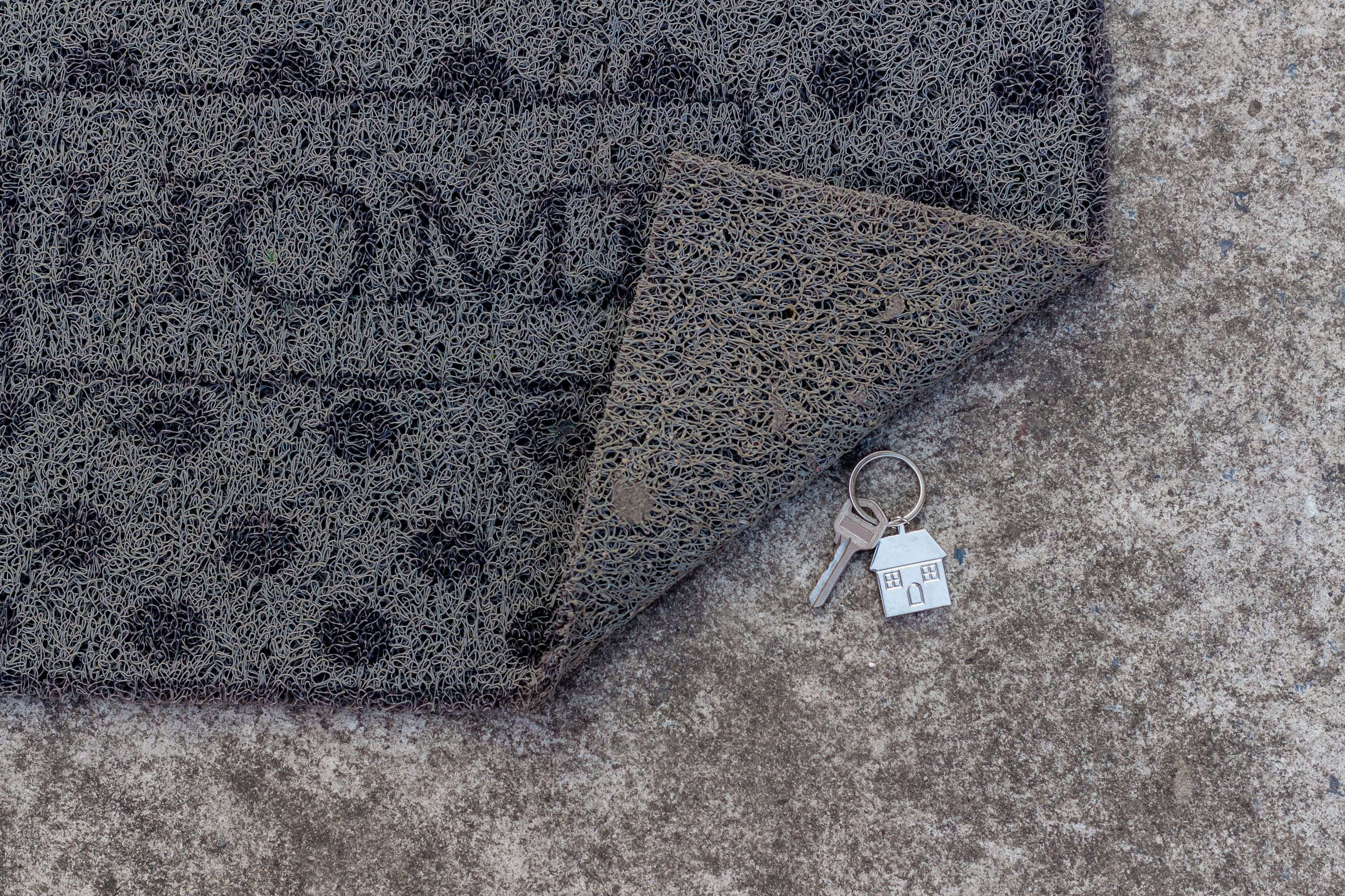 Doormat with the word "HOME" partially lifted, revealing two keys and a small house keychain underneath