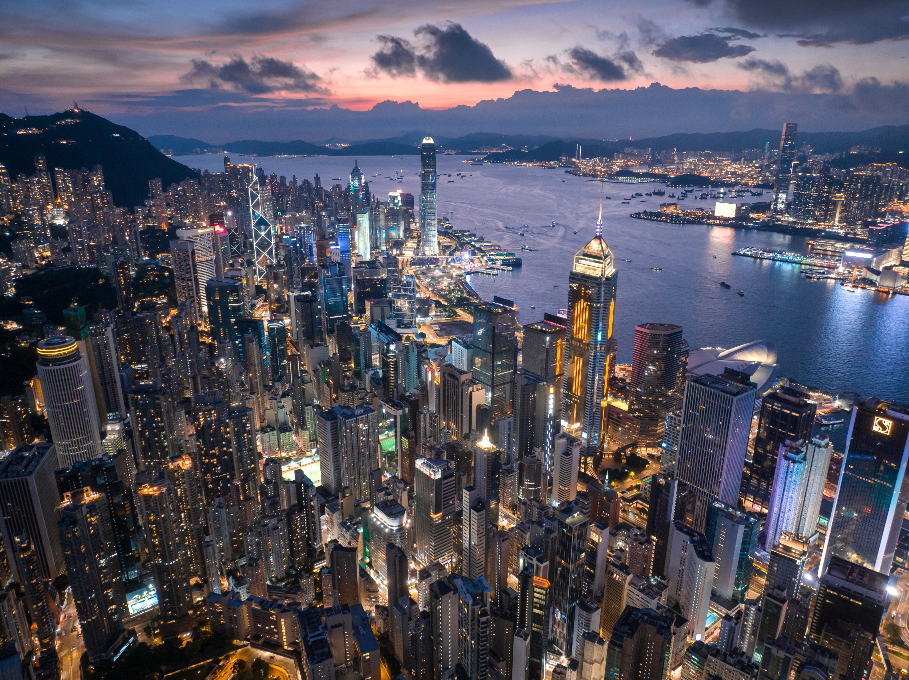 Aerial view of Hong Kong's illuminated skyline at dusk, showcasing numerous skyscrapers and a harbor, highlighting the city's bustling ambiance