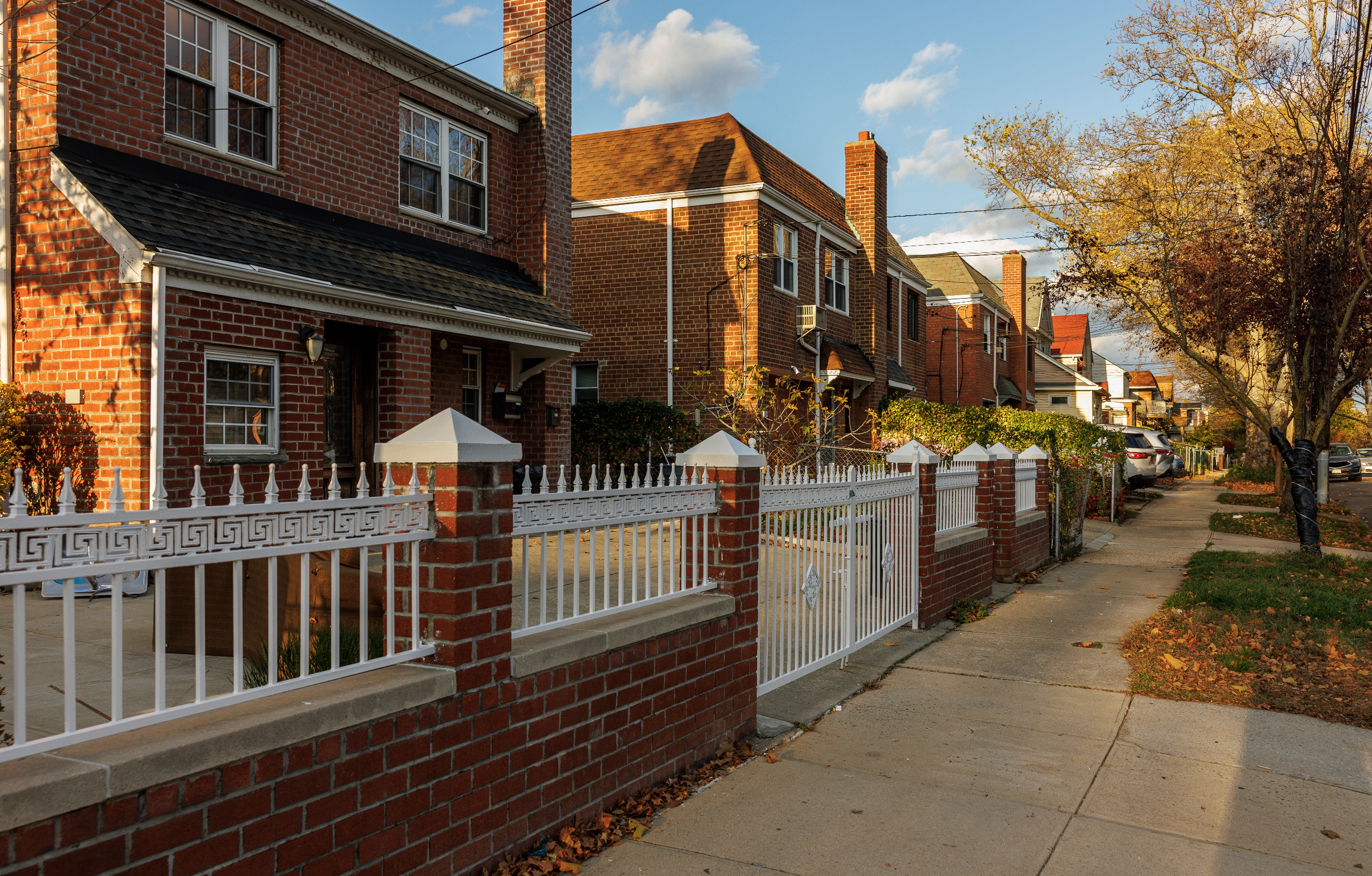 A residential street with brick houses and sidewalks lined with trees, under a partly cloudy sky