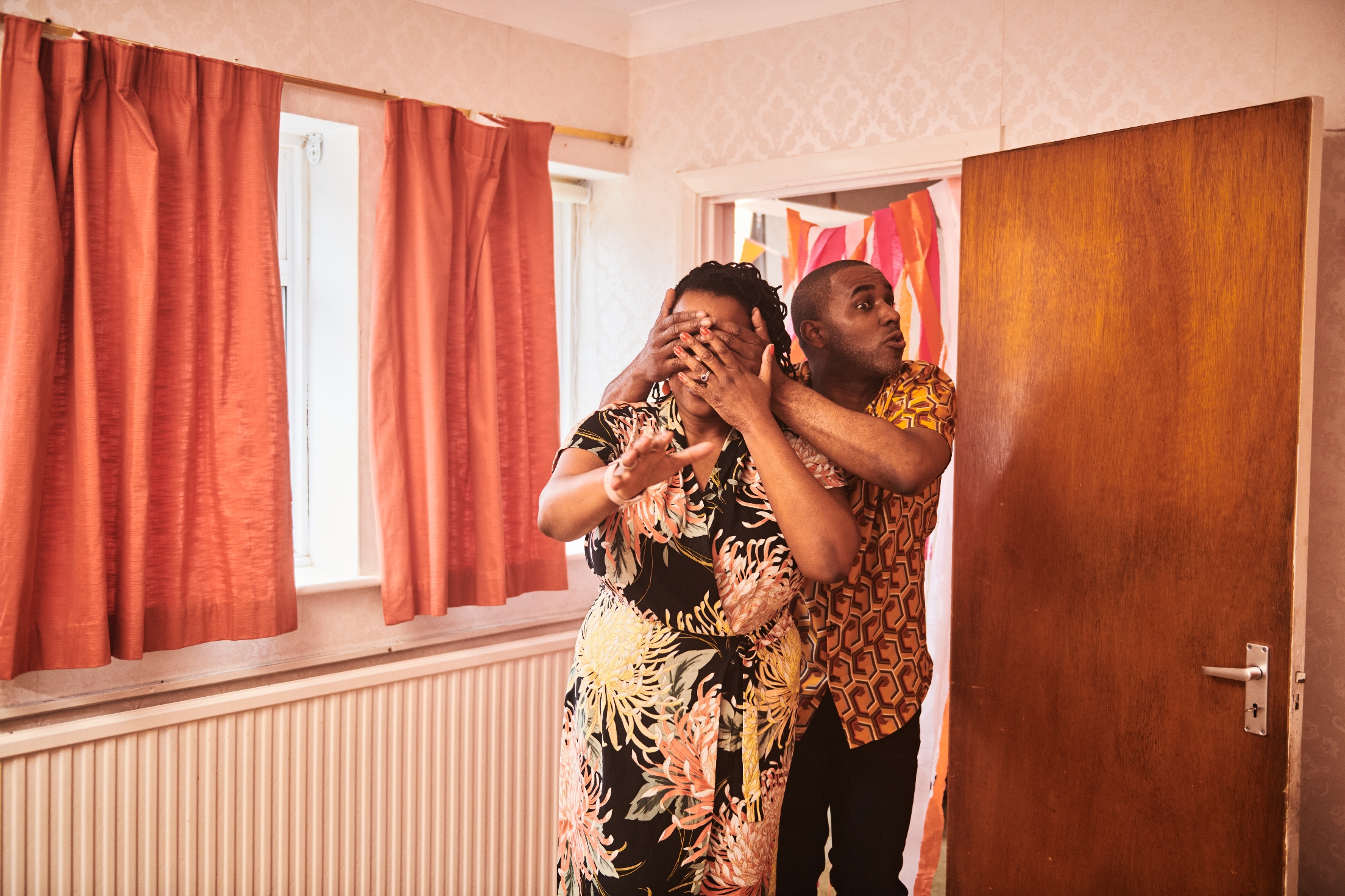 A person playfully covers another's eyes from behind in a cozy room, both smiling and wearing casual patterned outfits