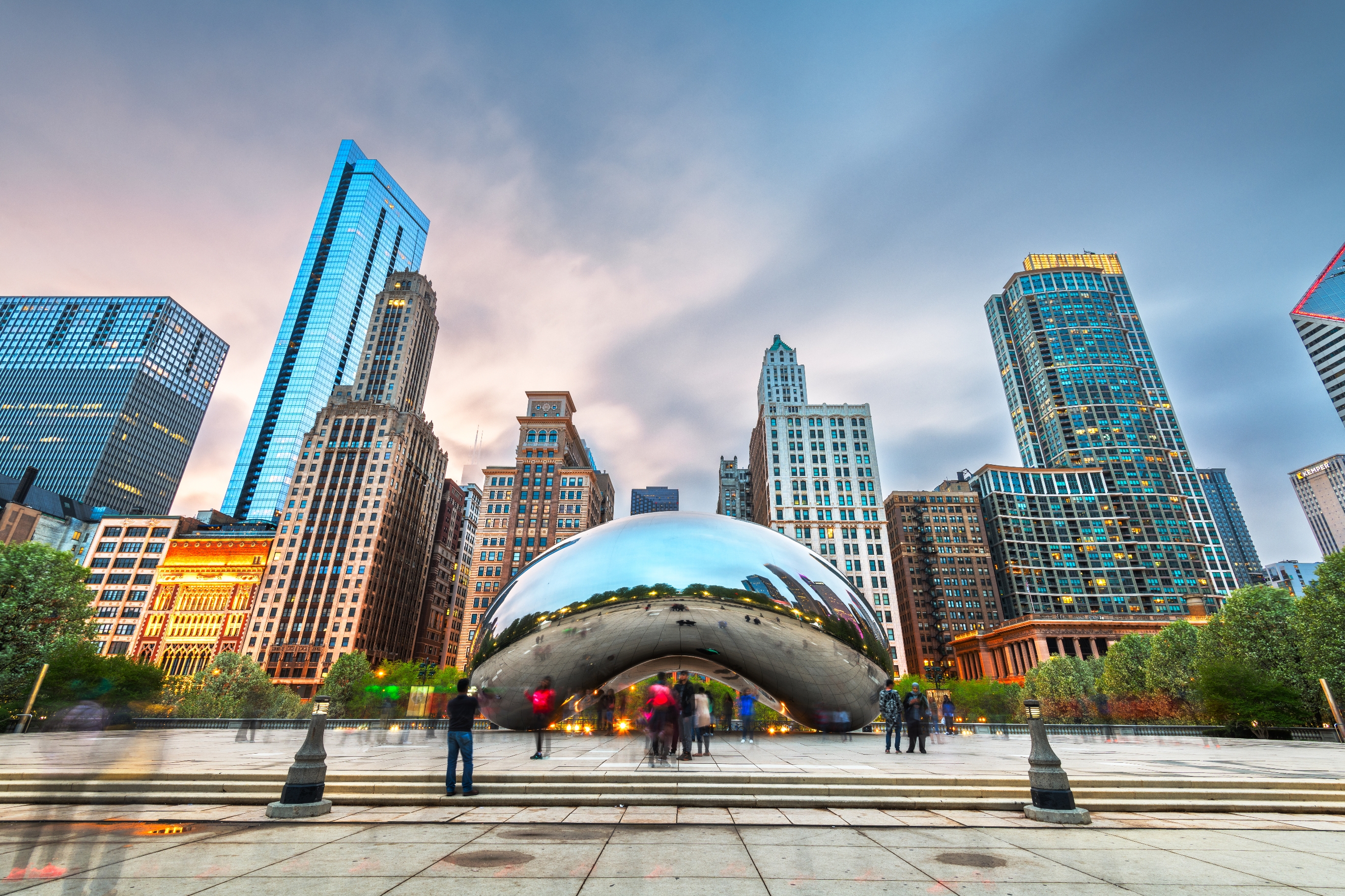People visit Chicago's Cloud Gate, a reflective bean-shaped sculpture, with a skyline of tall buildings in the background