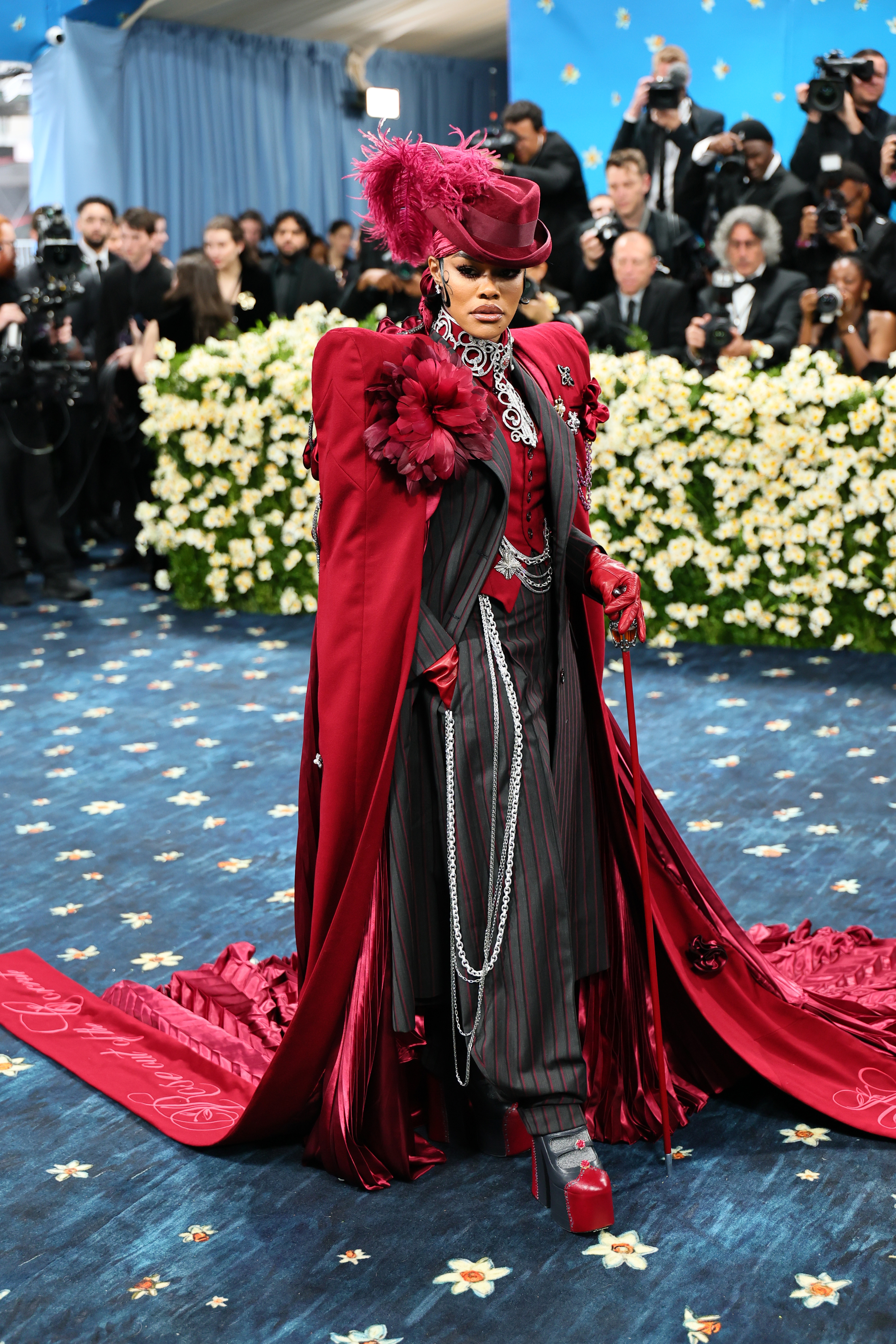 Person wearing elaborate Victorian-inspired outfit with red cape, feathered hat, and striped suit on a floral-themed event carpet