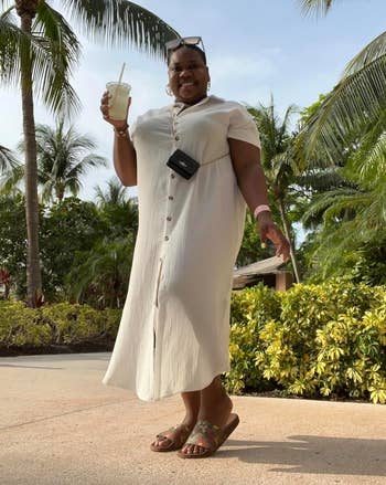 Person outdoors successful a button-up formal holds a drink, smiling nether thenar trees. They deterioration sunglasses and sandals, with a tiny crossbody bag